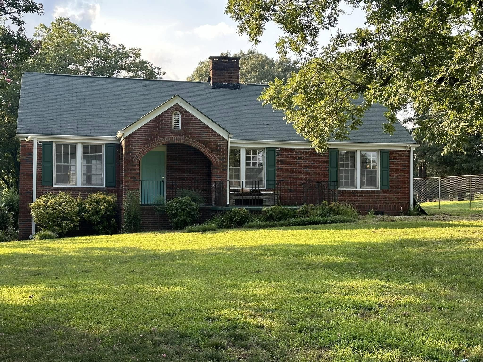 Brick house with a blue roof, green shutters, and a small front porch with steps, surrounded by a green lawn and trees.