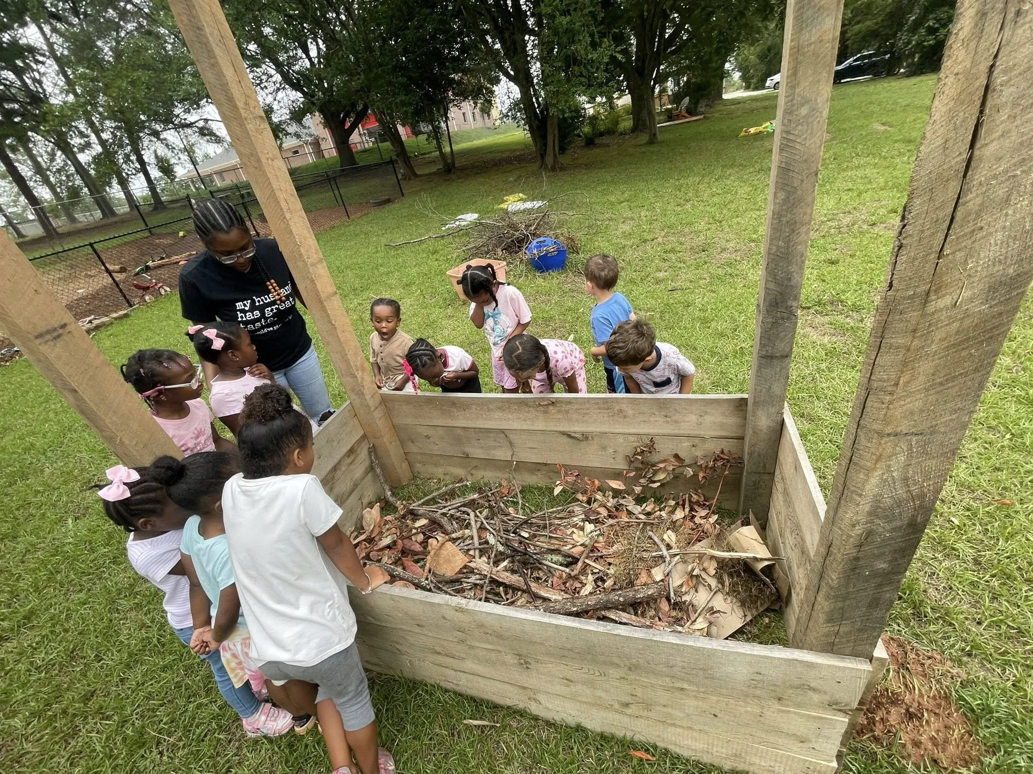 Group of children and an adult gathered around a small outdoor compost bin filled with leaves and branches.