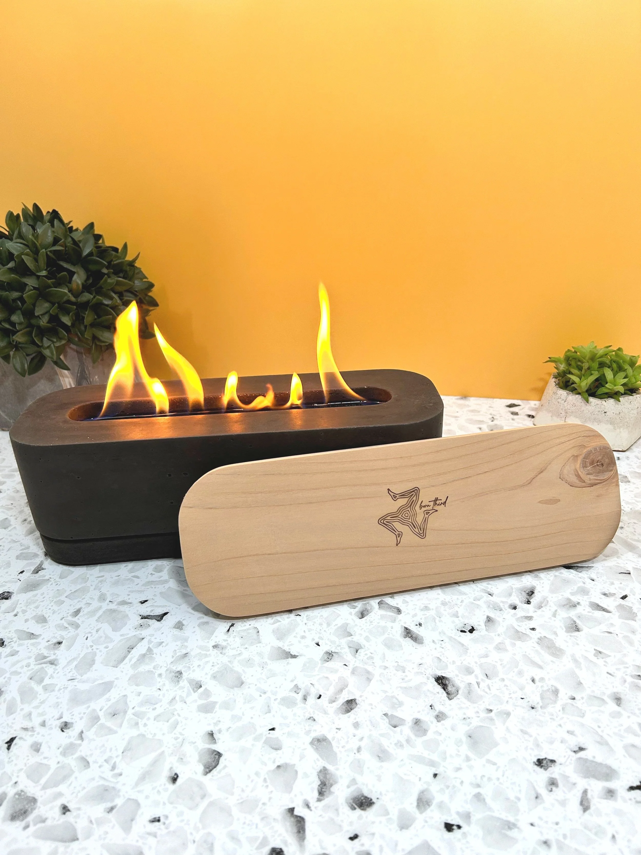 A modern tabletop fireplace with flames, a wooden lid, and two small potted succulents on a white speckled surface against a yellow wall.