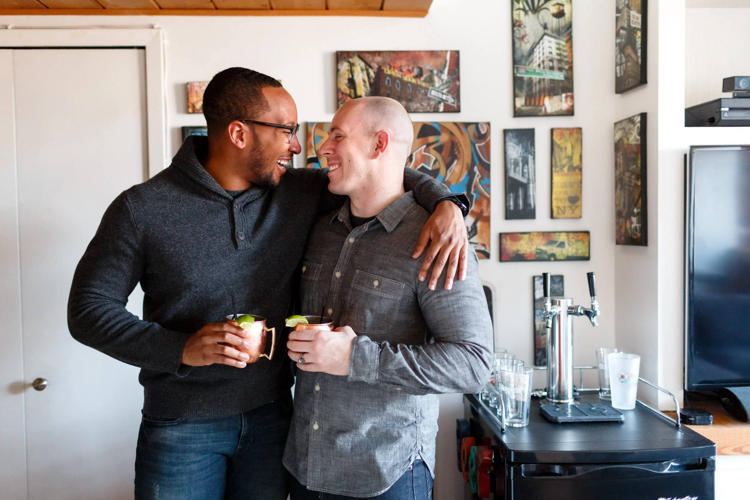 A couple with their arms around each other each holding a drink and smiling in their home 