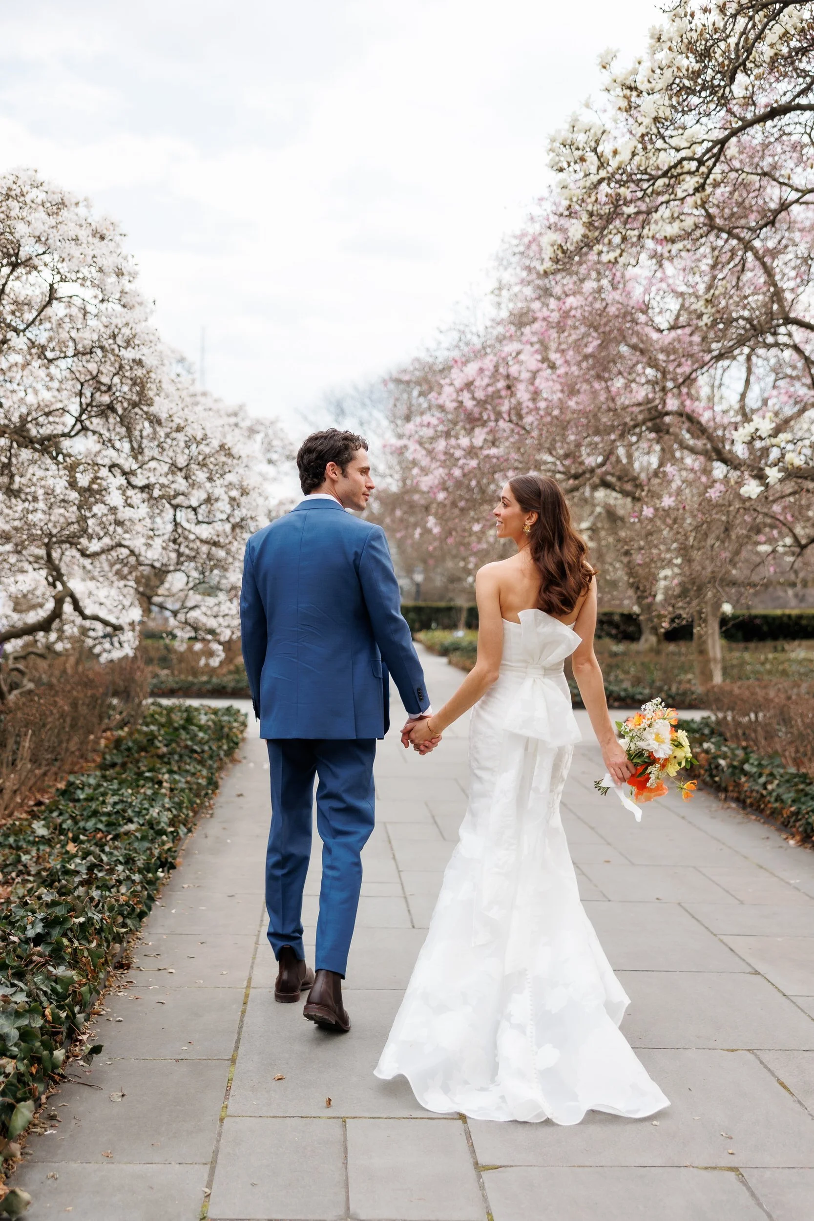 Newlyweds holding hands and smiling at each other as they walk through a park 