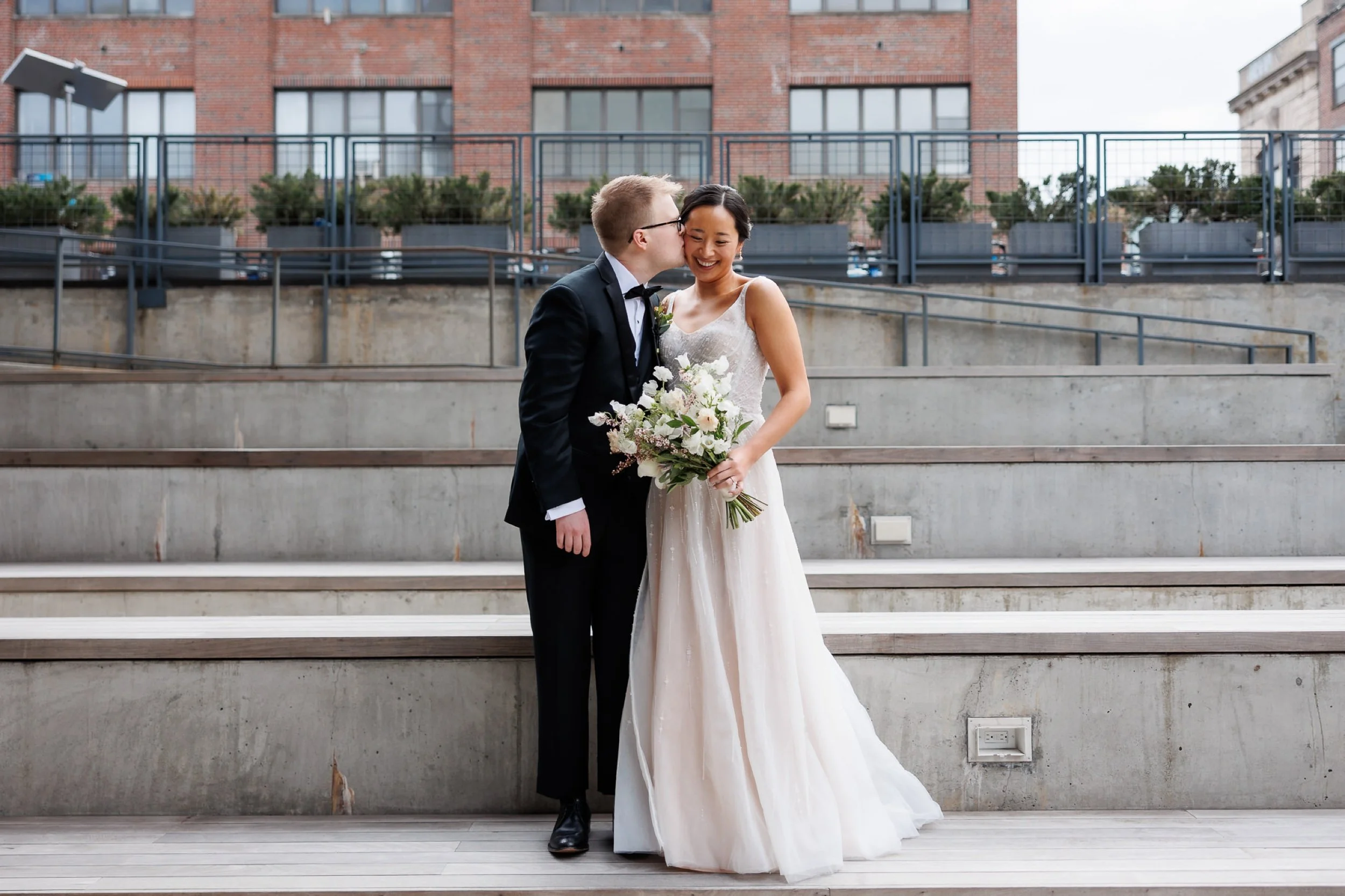 Man in black suit kisses woman in blush wedding dress on the cheek