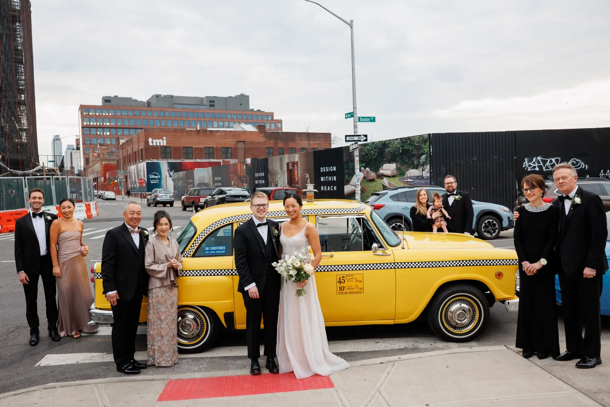 Couple in wedding attire poses with their families in front of a vintage NYC cab
