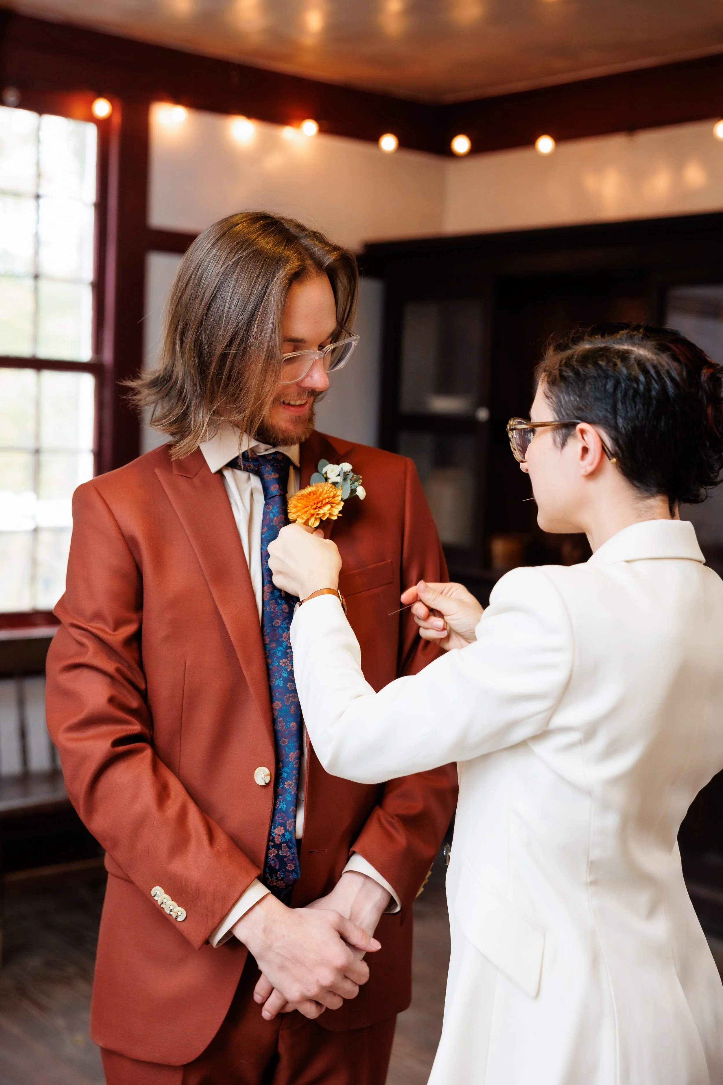 Couple gets ready together before their wedding as one partner pins on the boutonniere for their partner at Ashokan Center