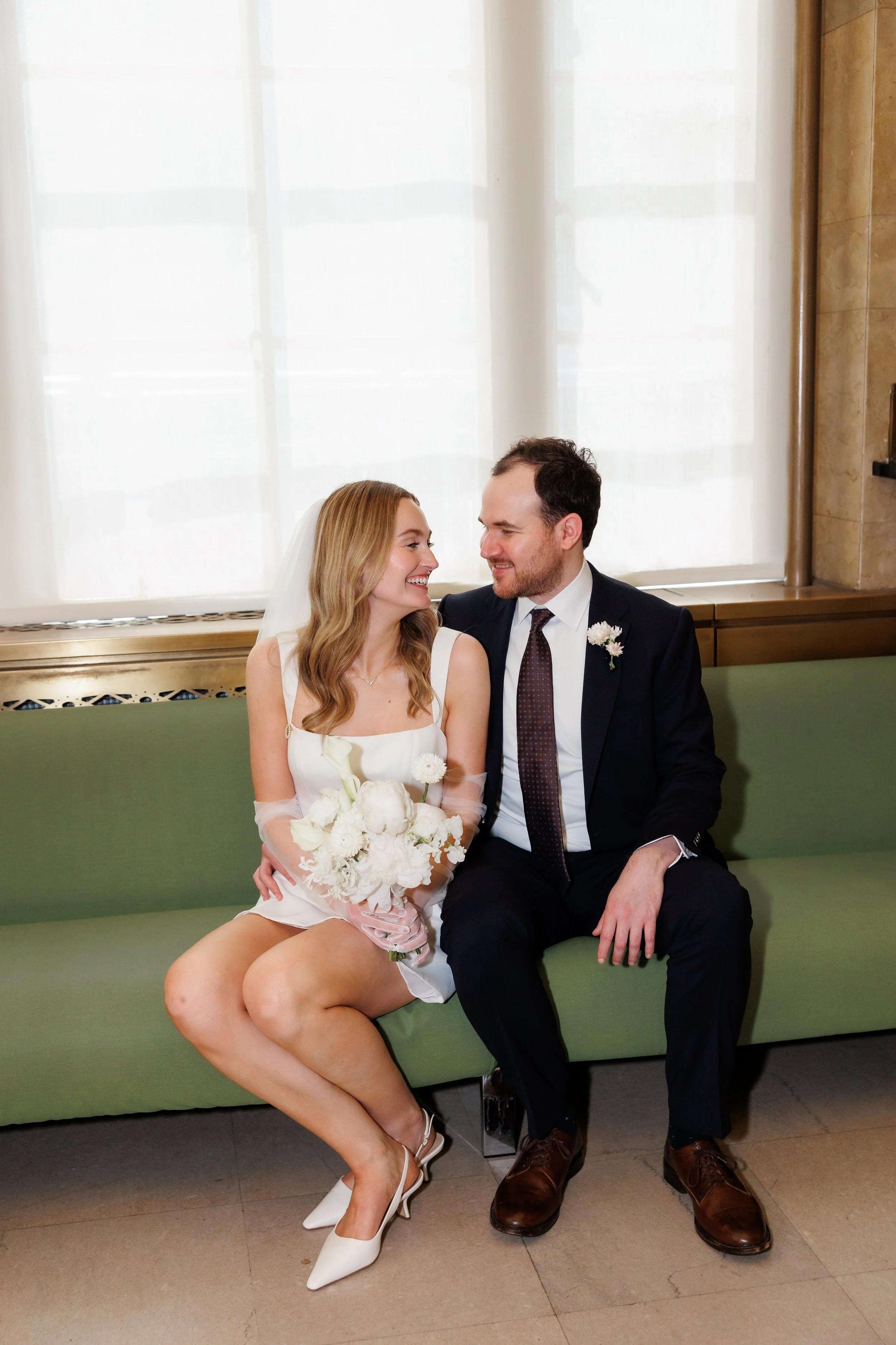 Couple cuddles on the green couches at the NYC marriage bureau before their elopement ceremony