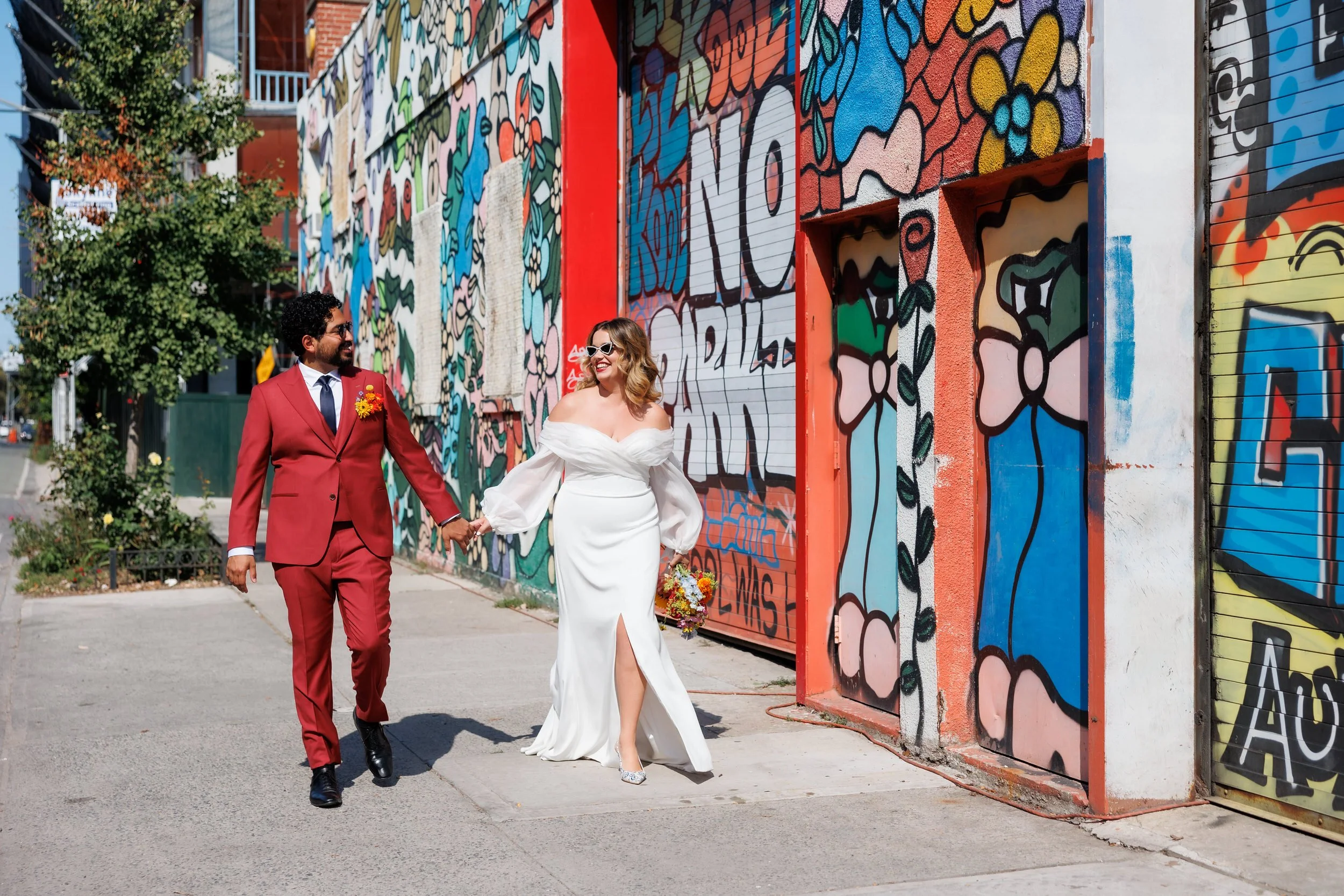 A newlywed couple holding hands and walking on a sidewalk next to a colorful mural