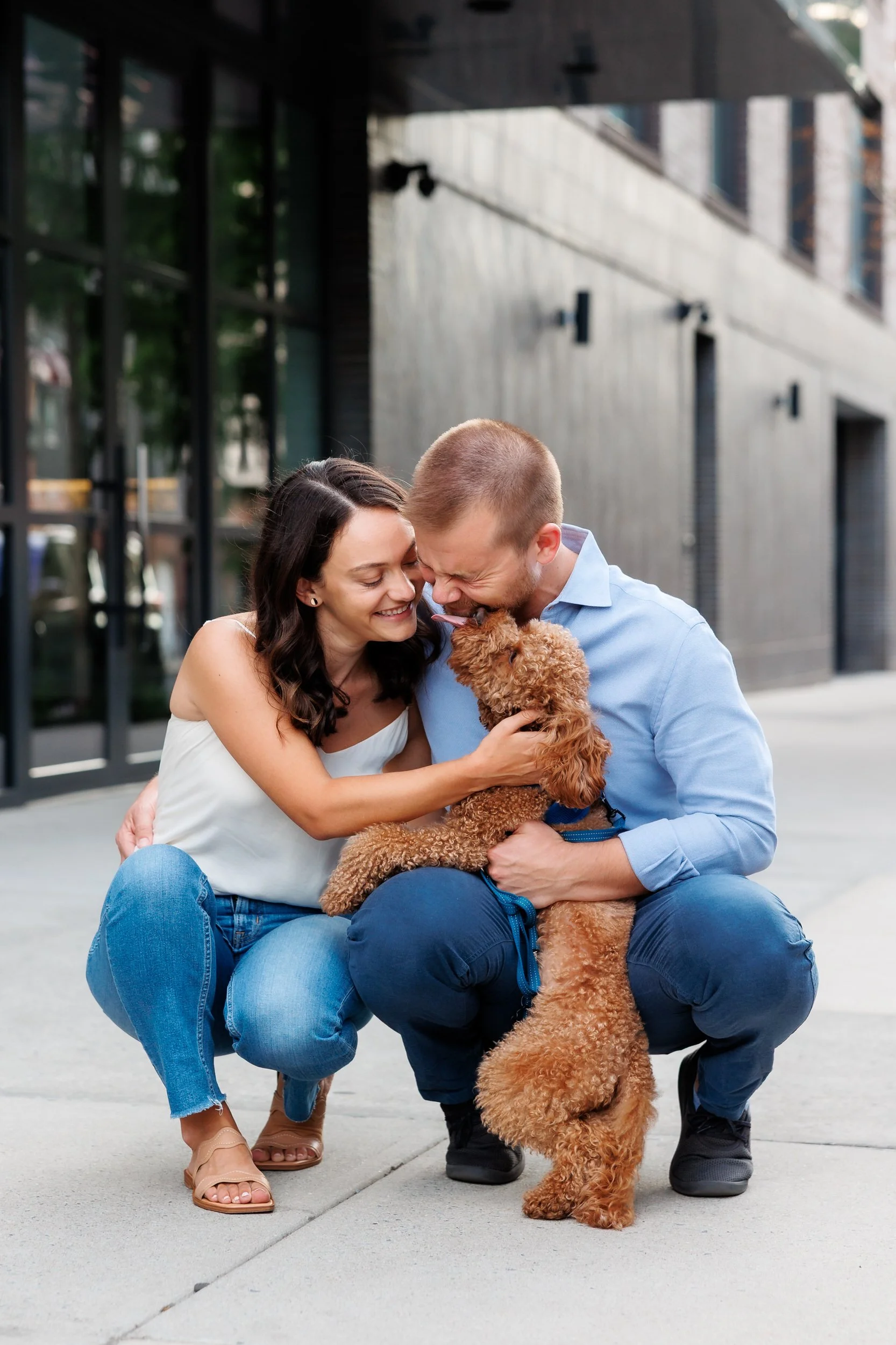 A couple leaning down and petting as dog as it licks one of their faces 