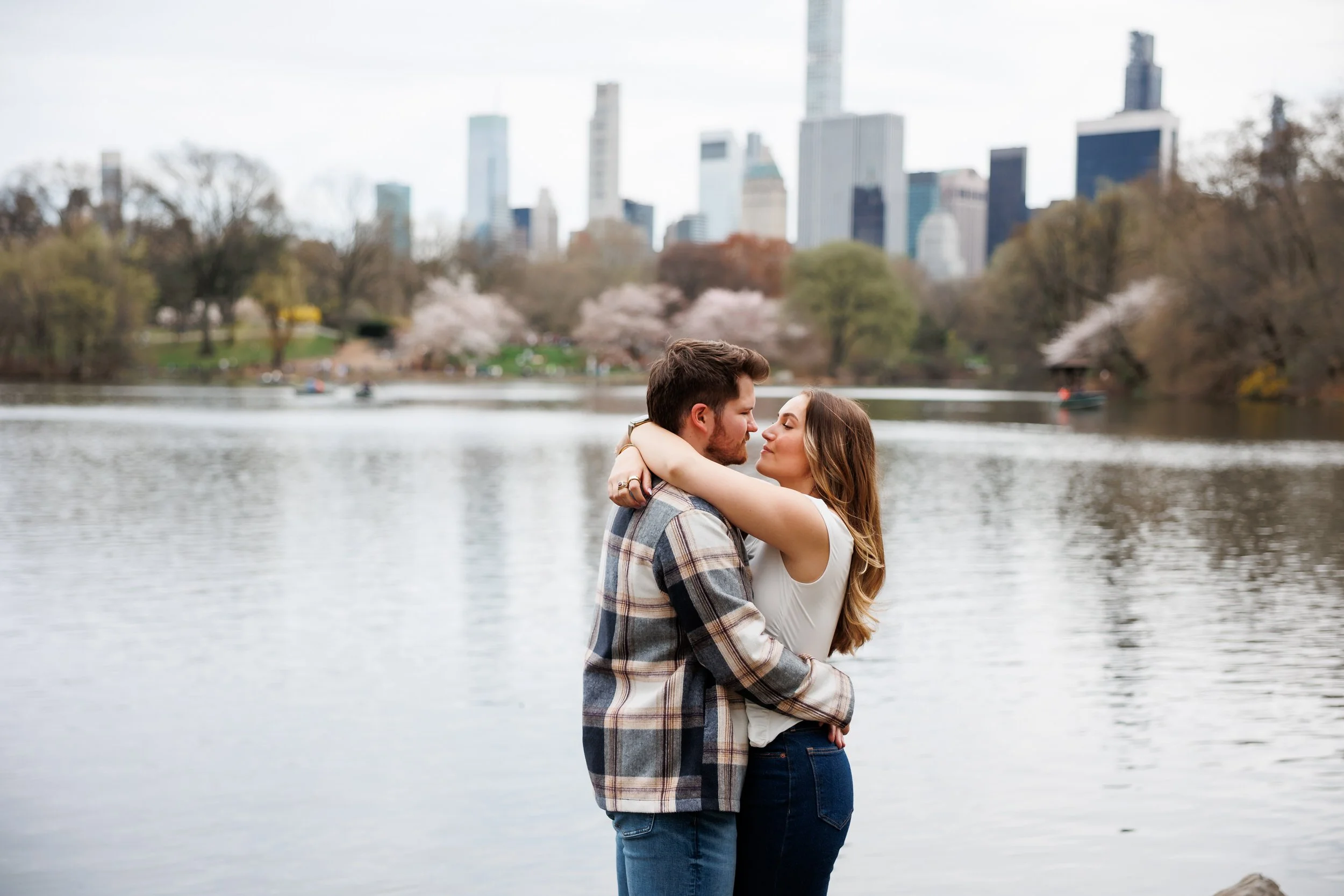 A couple with their arms around each other about to kiss by a lake with a city skyline in the distance 