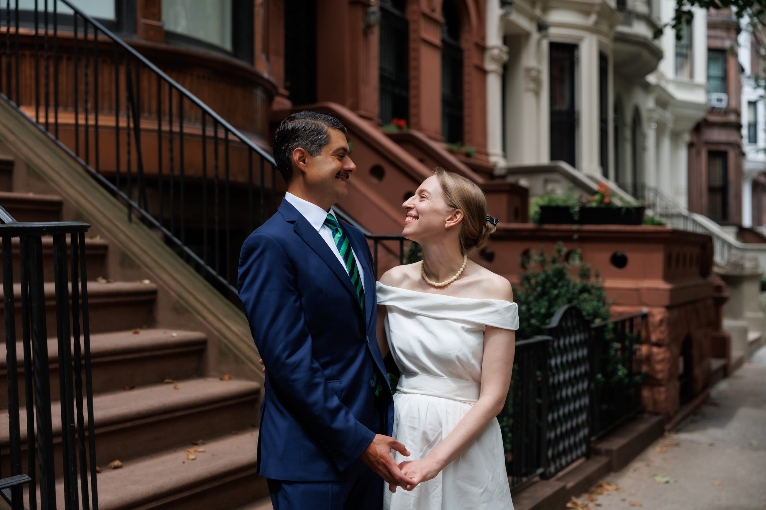Newlyweds holding hands and smiling at each other while standing next to brownstones