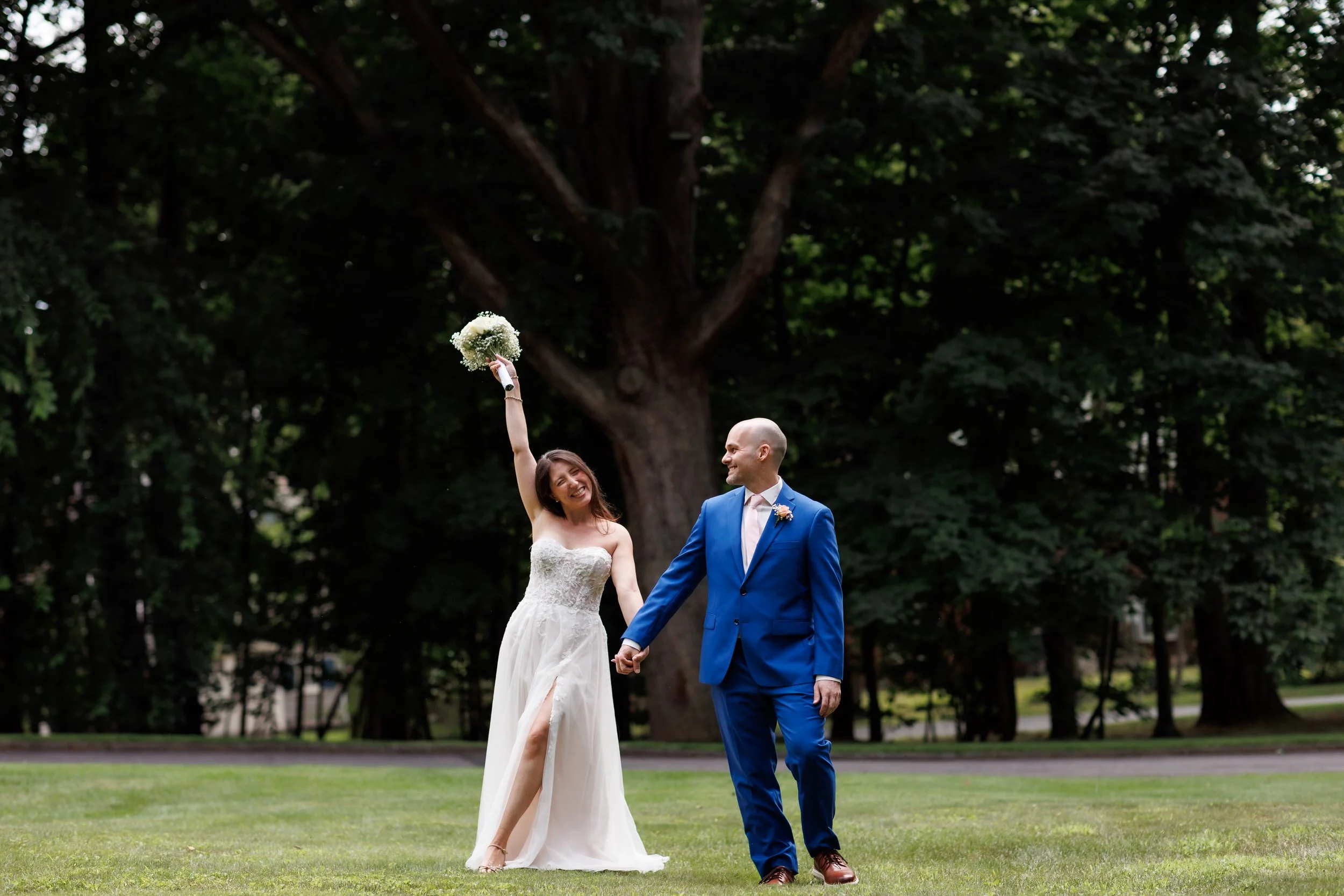 Couple walks towards the camera in wedding attire, and bride raises her bouquet in the air in celebration