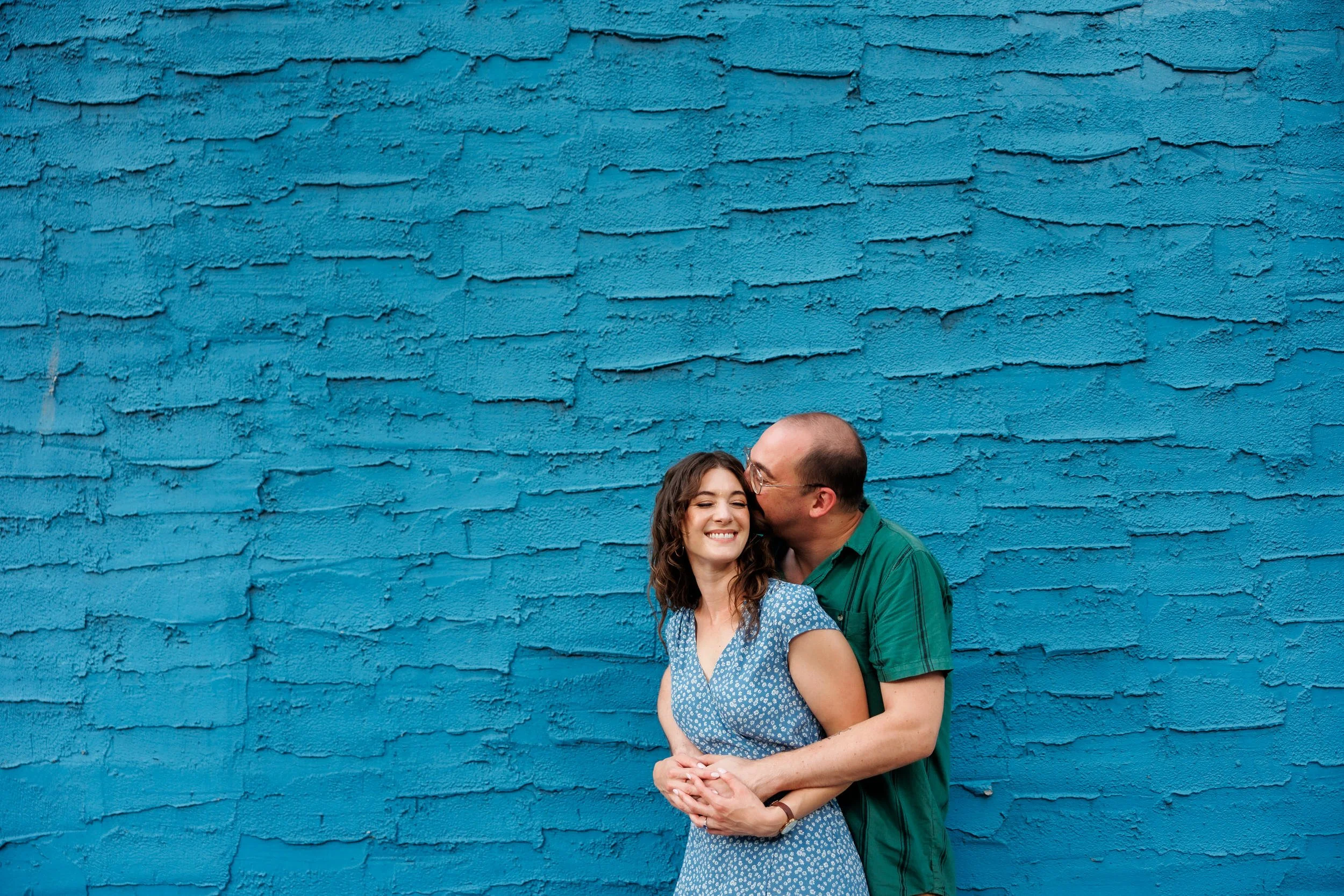 A person standing behind their partner wrapping them in a hug and kissing their head