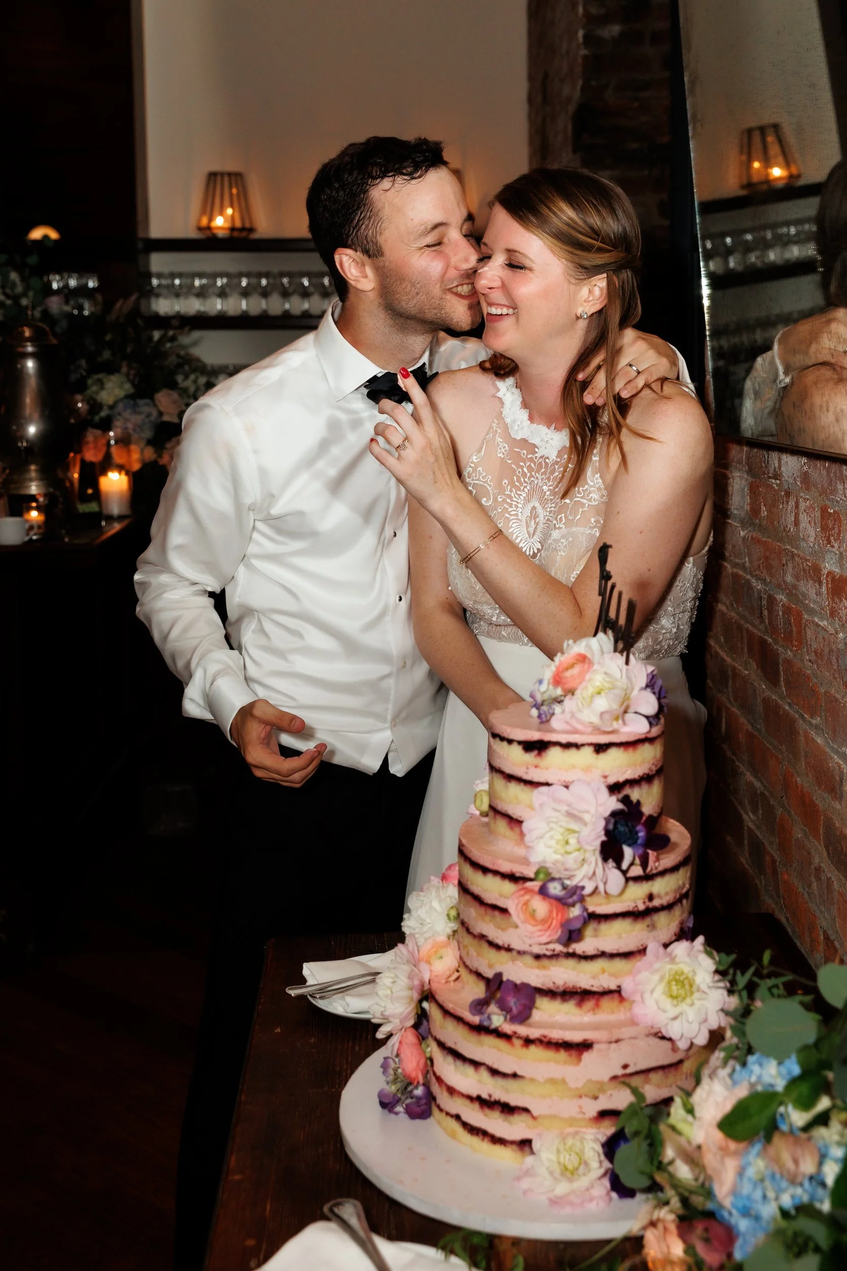 A person leaning in to kiss their partner's cheek as they cut their wedding cake 