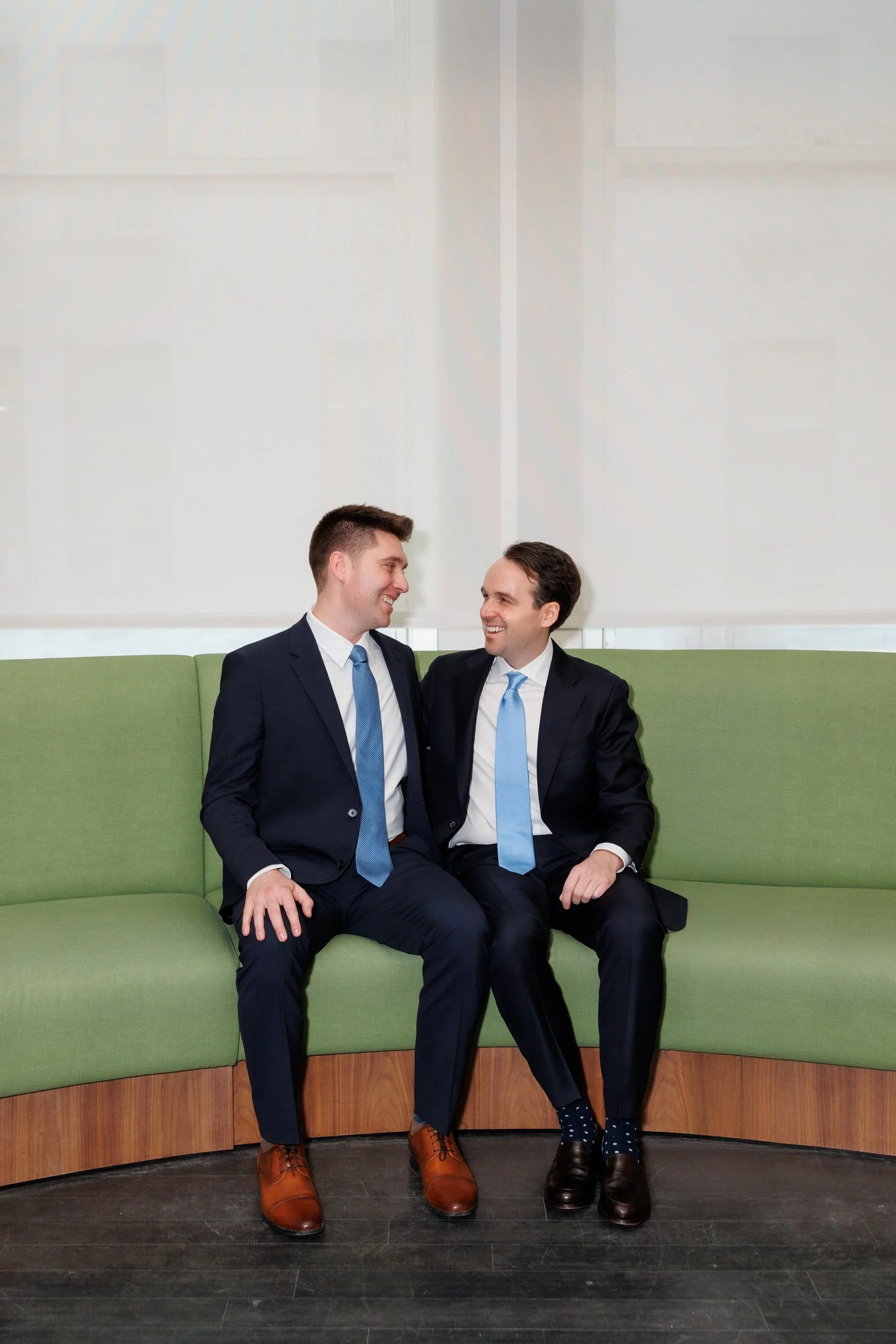 Two newlyweds in suits smiling at each other as they sit on a green couch