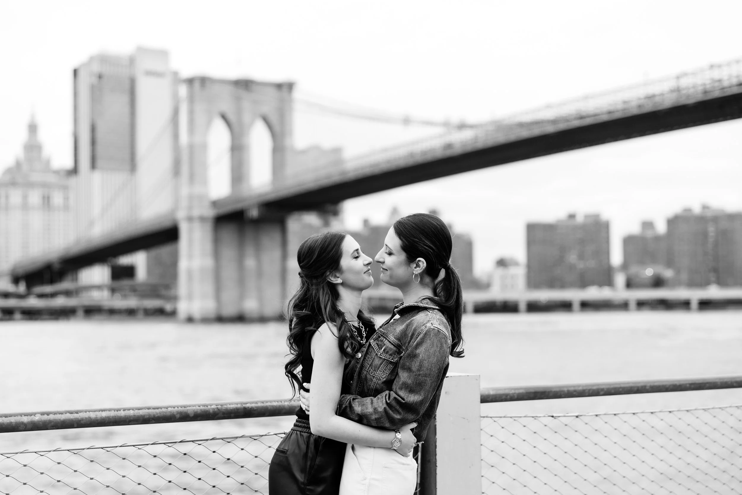 A couple leaning in to kiss in front of a bridge with a city skyline behind them 