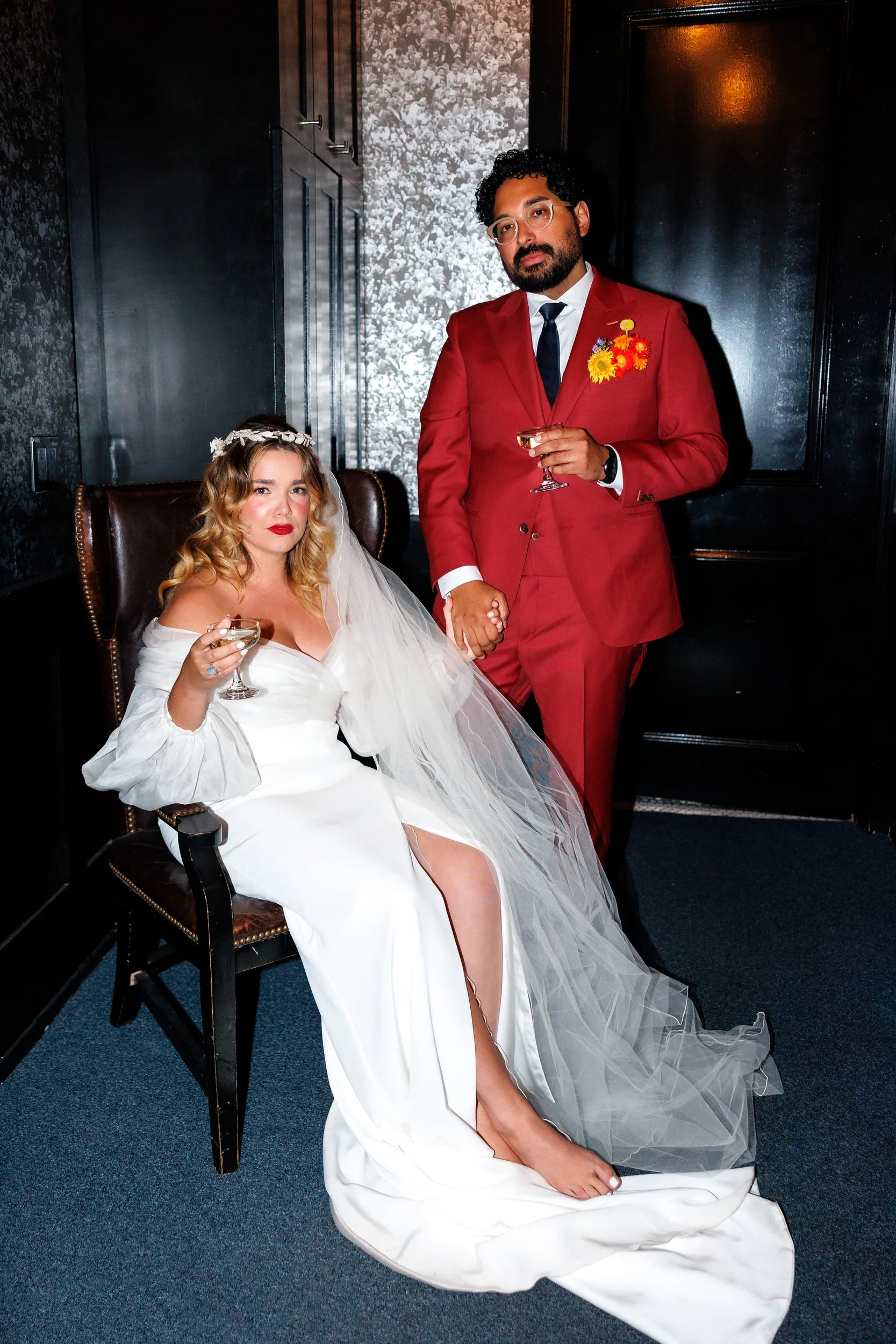 A newlywed sitting in a chair holding a drink while their partner stands next to them holding their other hand