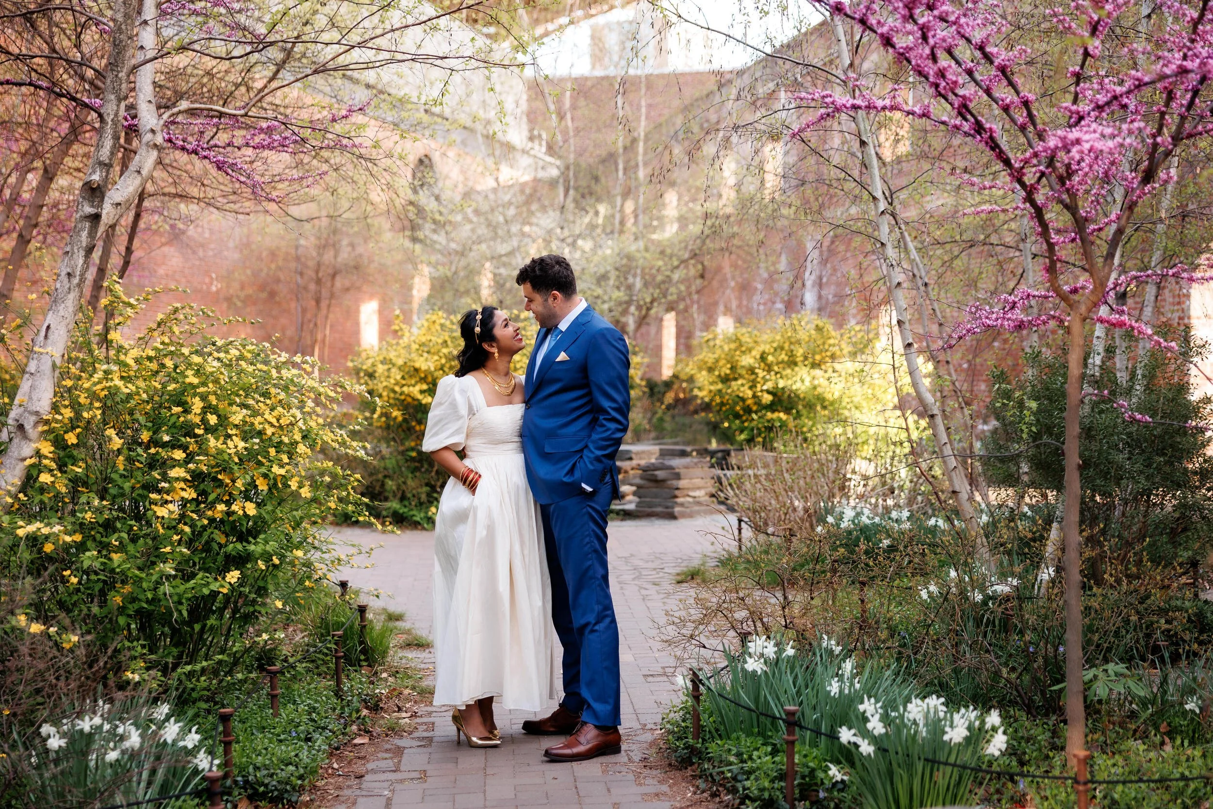 A newlywed couple standing close together smiling in a colorful garden 