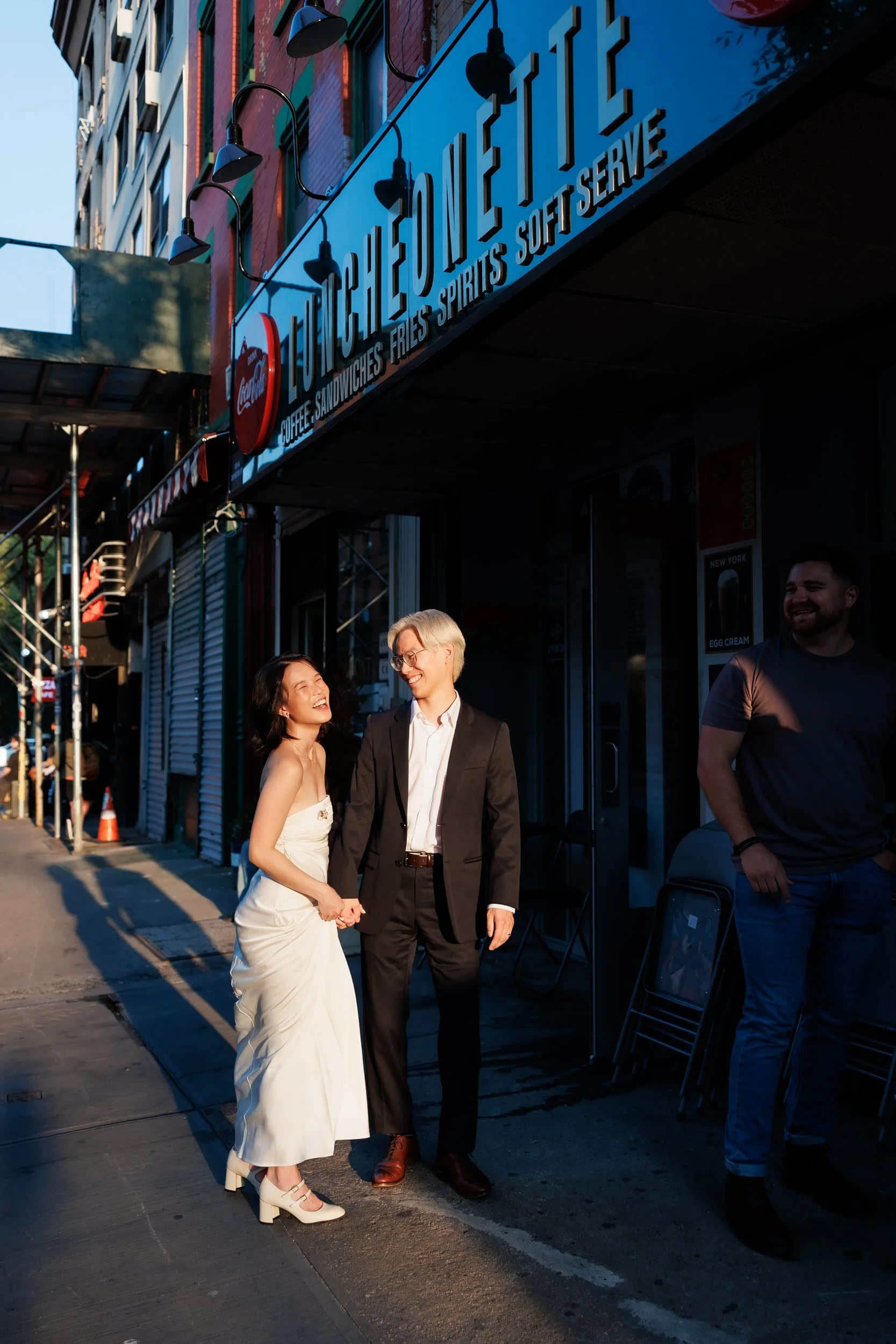 A couple holding hands and laughing outside of a building on a shopping street. 