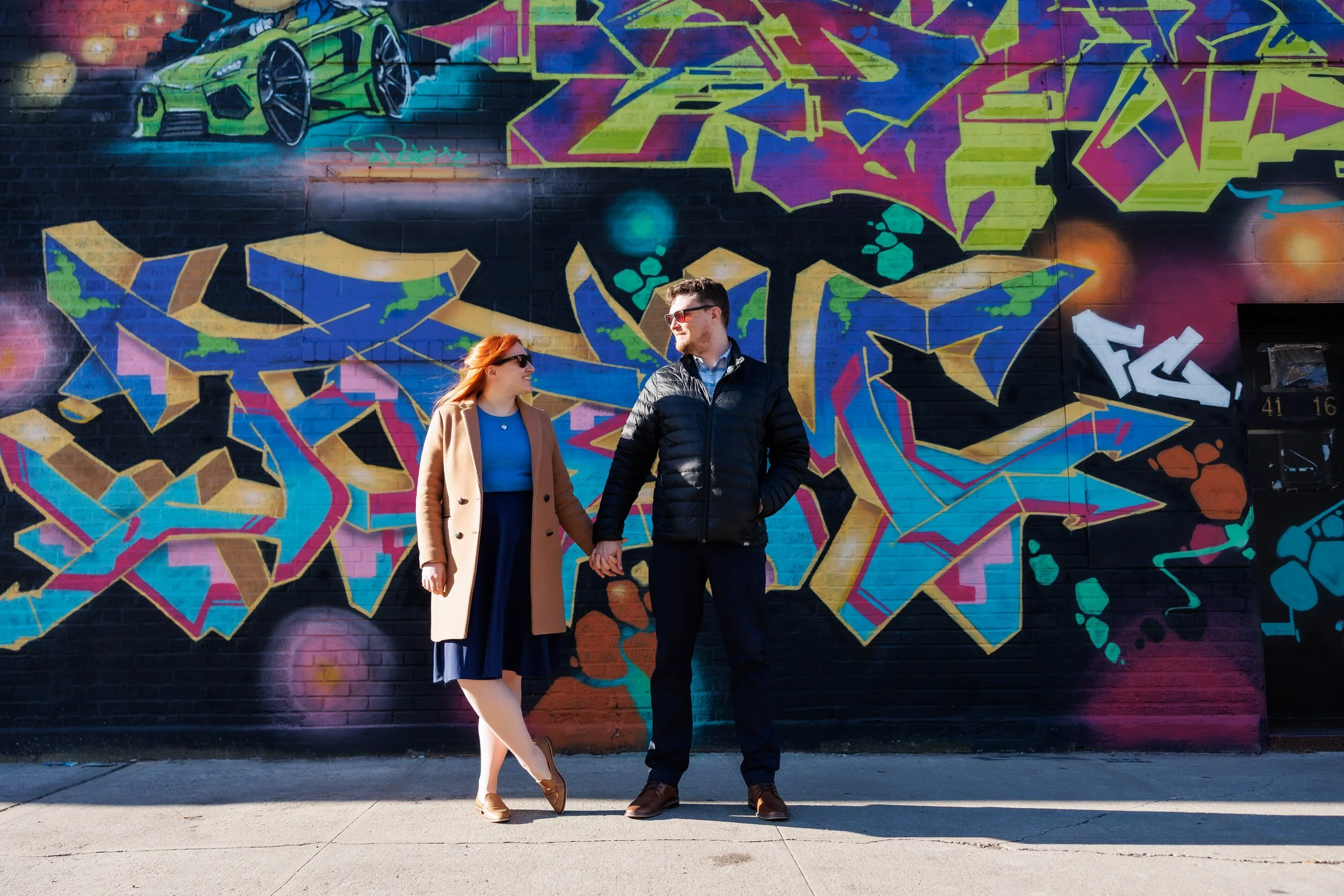 A couple holding hands and looking at each other with colorful graffiti on the wall behind them 