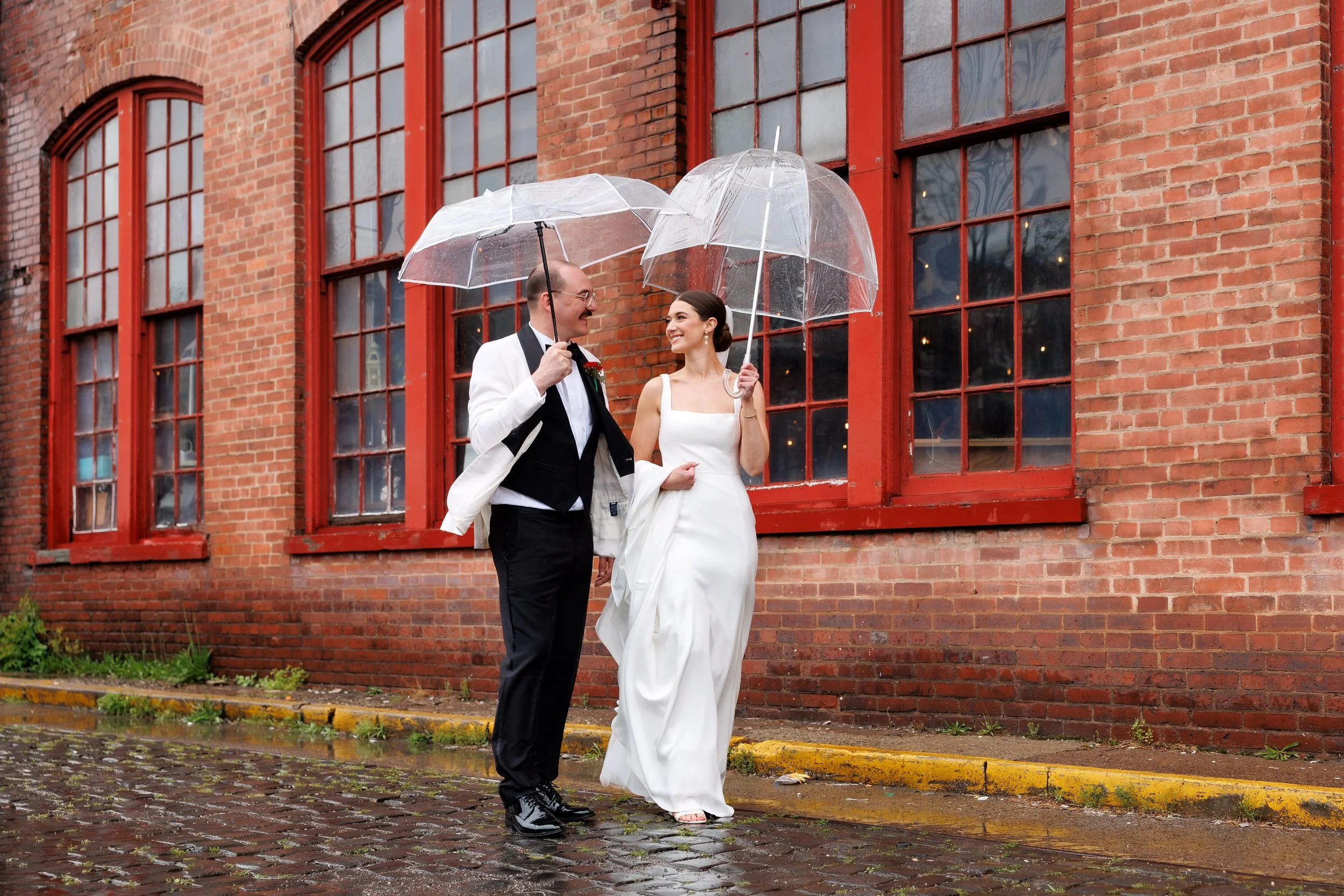 Newlyweds holding hands and walking along a cobblestone road as they each hold clear umbrellas 