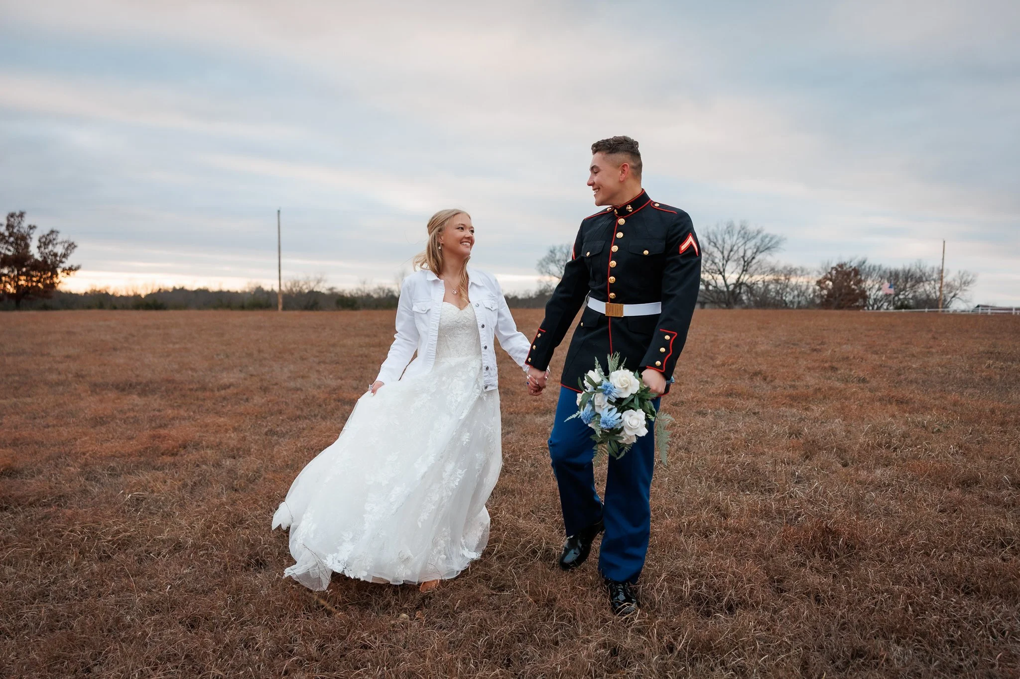 couple walks through field in wedding attire at sunset