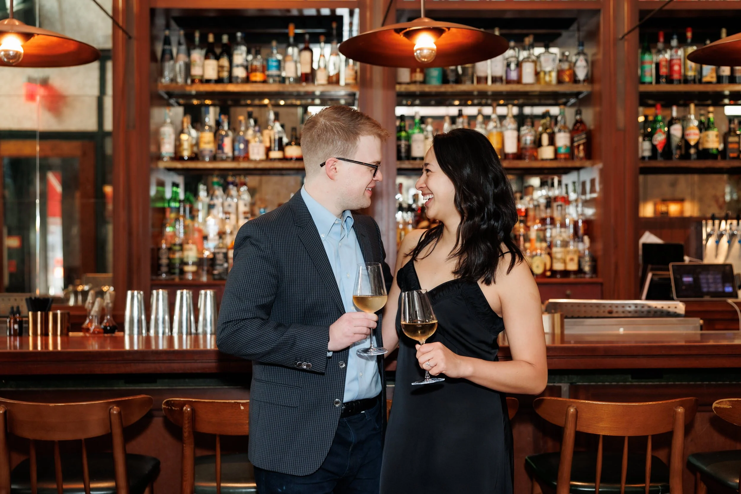 A couple holding wine glasses at a bar while smiling at each other 