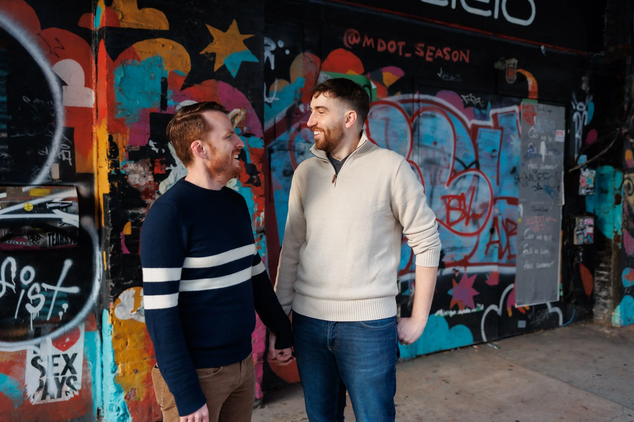 A couple holding hands and smiling at each other in front of a wall covered in graffiti 