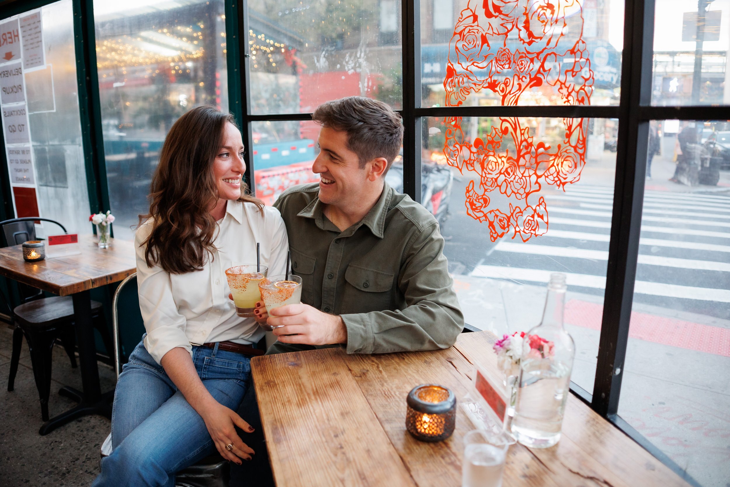 A couple sitting at a small table together while toasting two cocktails