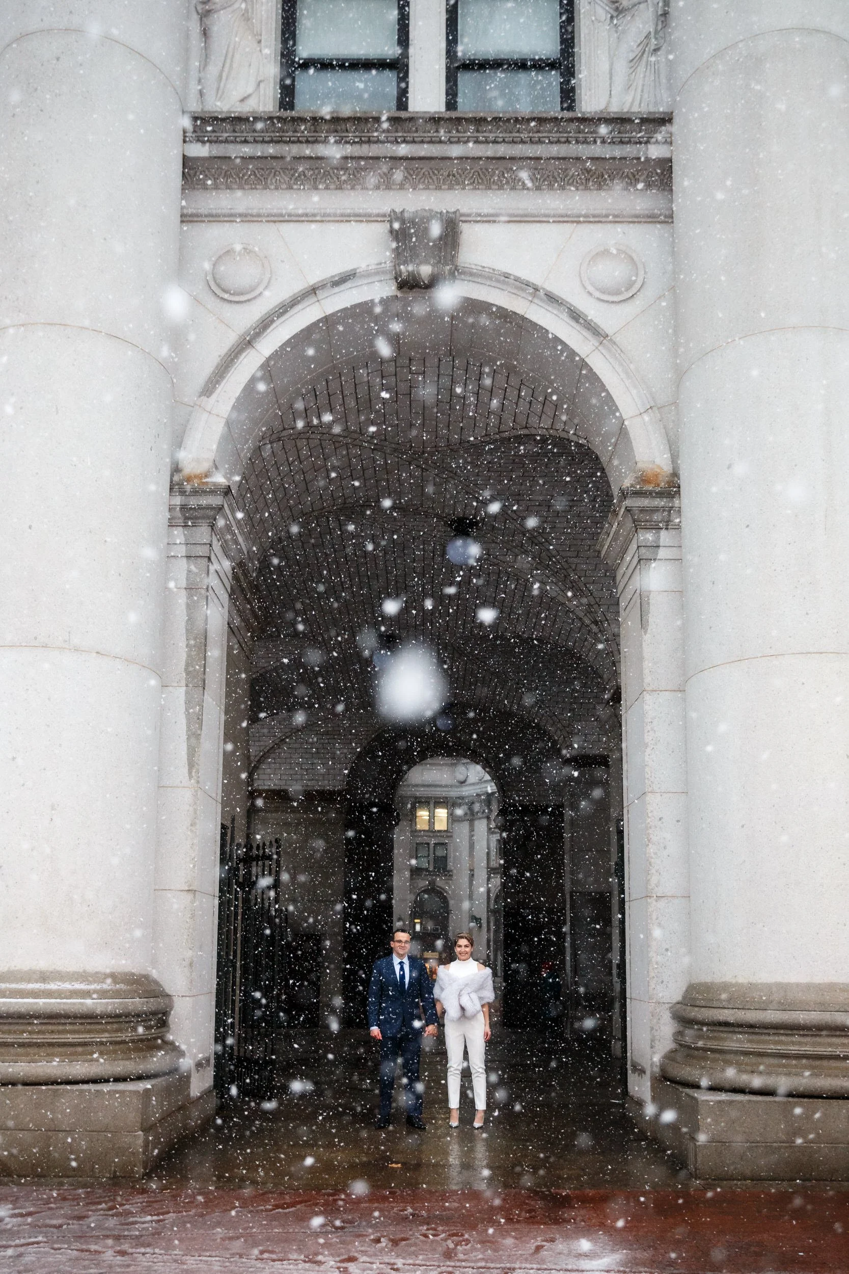 a wedding couple standing in an alcove as snow falls 