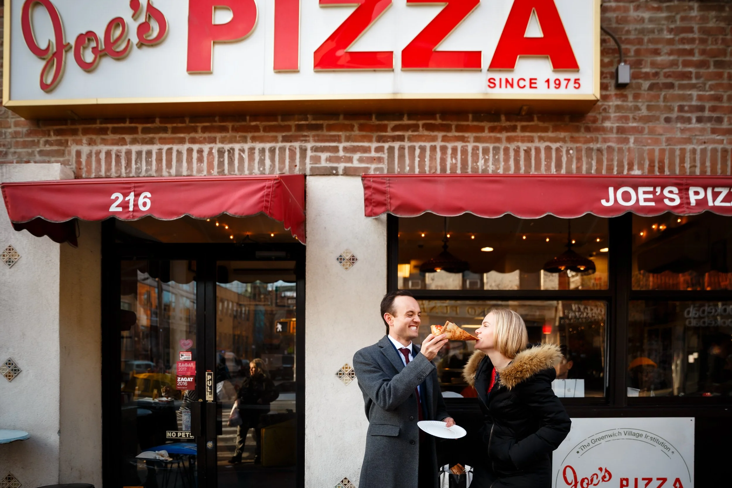 A person feeding their partner pizza outside of a pizzeria 