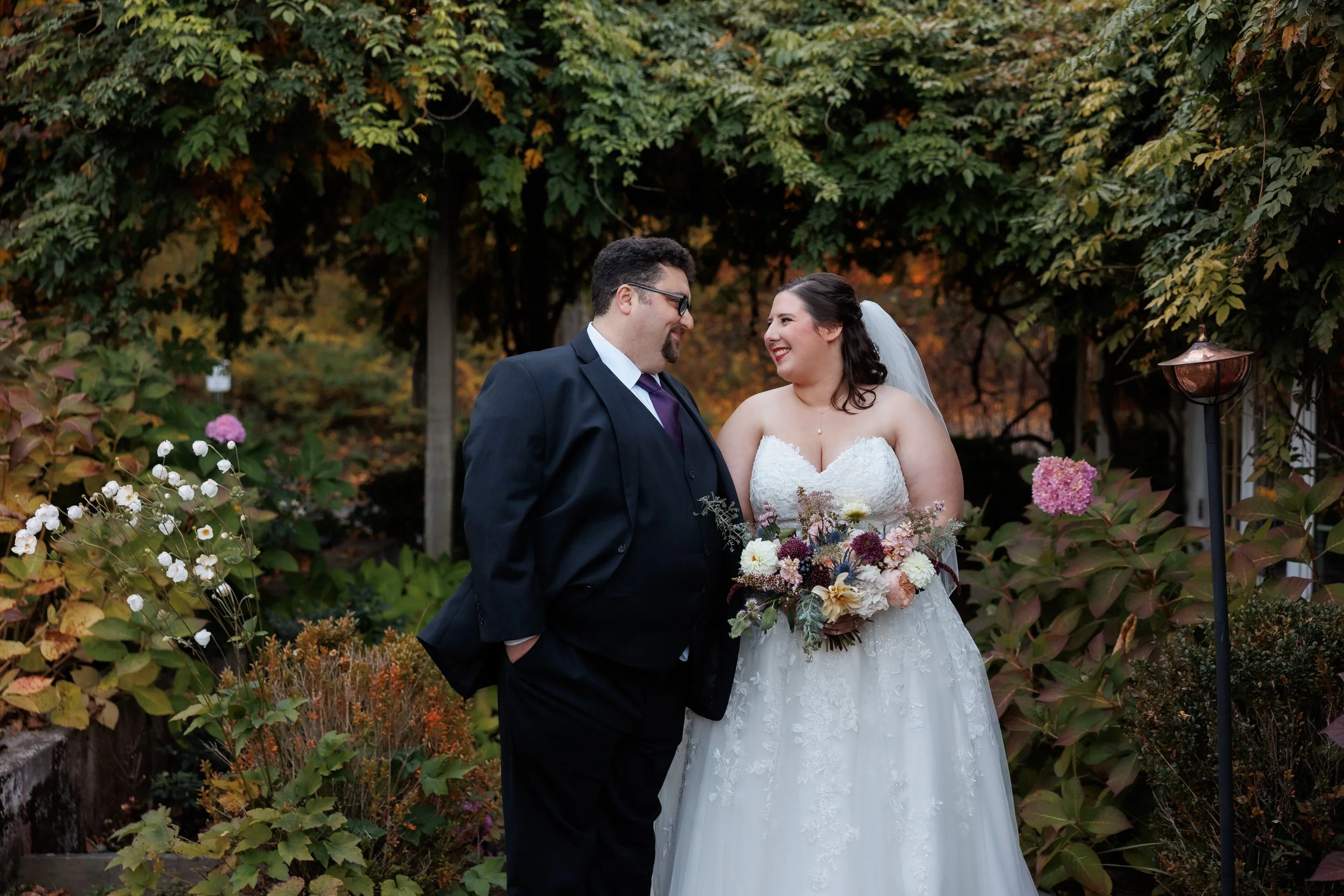 A newlywed couple standing close and smiling at each other while one holds a bouquet of flowers