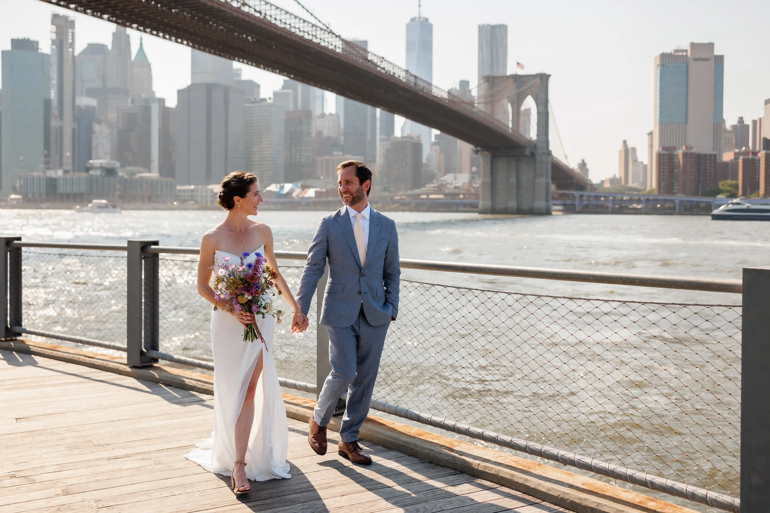 Newlyweds holding hands and walking along a riverside path with a city skyline in the background 
