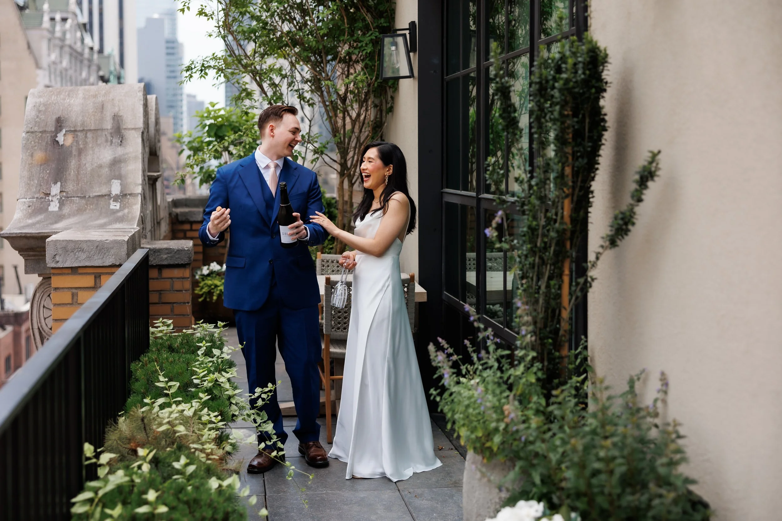 A wedding couple laughing as they pop a bottle of champagne on a balcony 