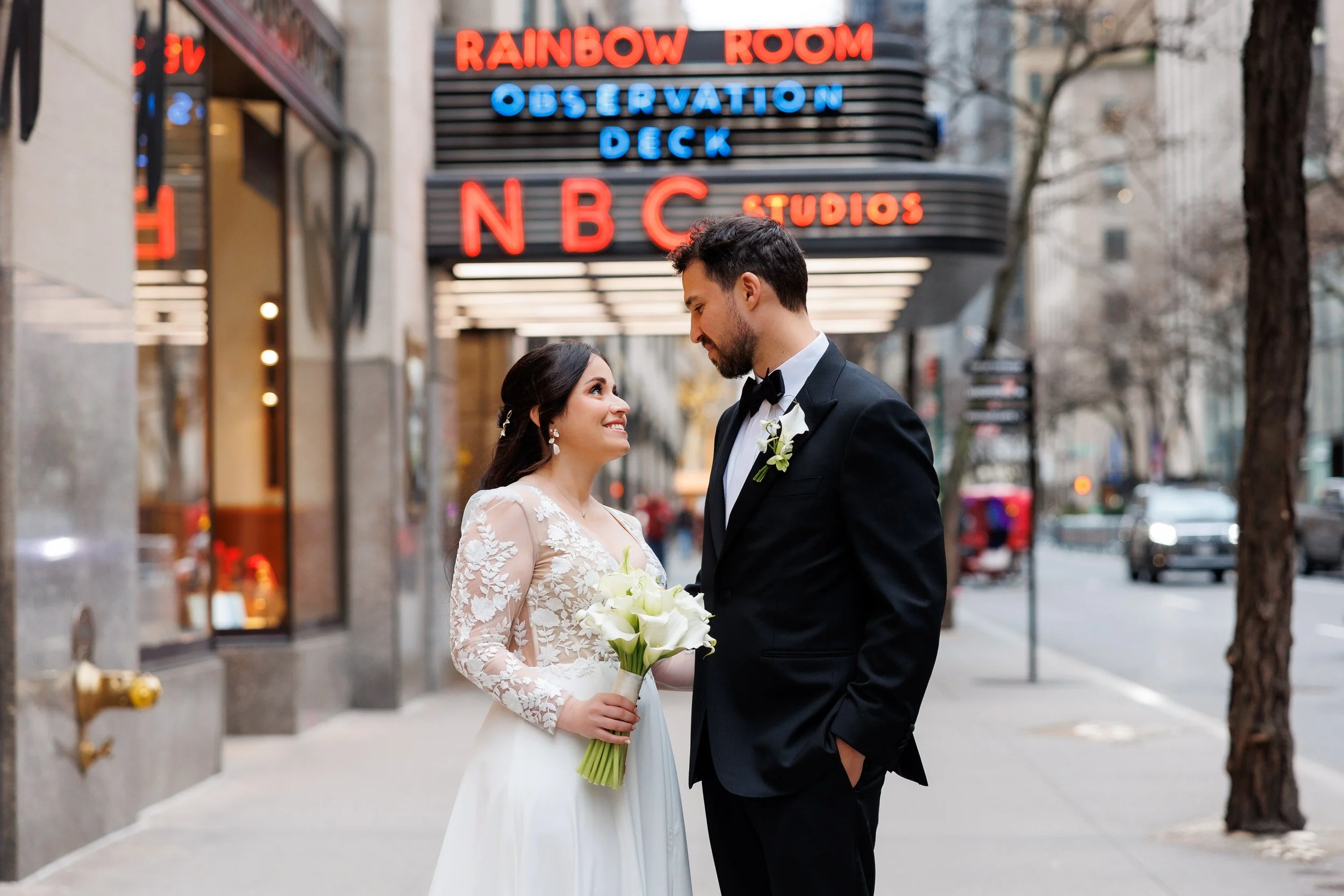 A newlywed couple standing on a city sidewalk as they look at each other lovingly