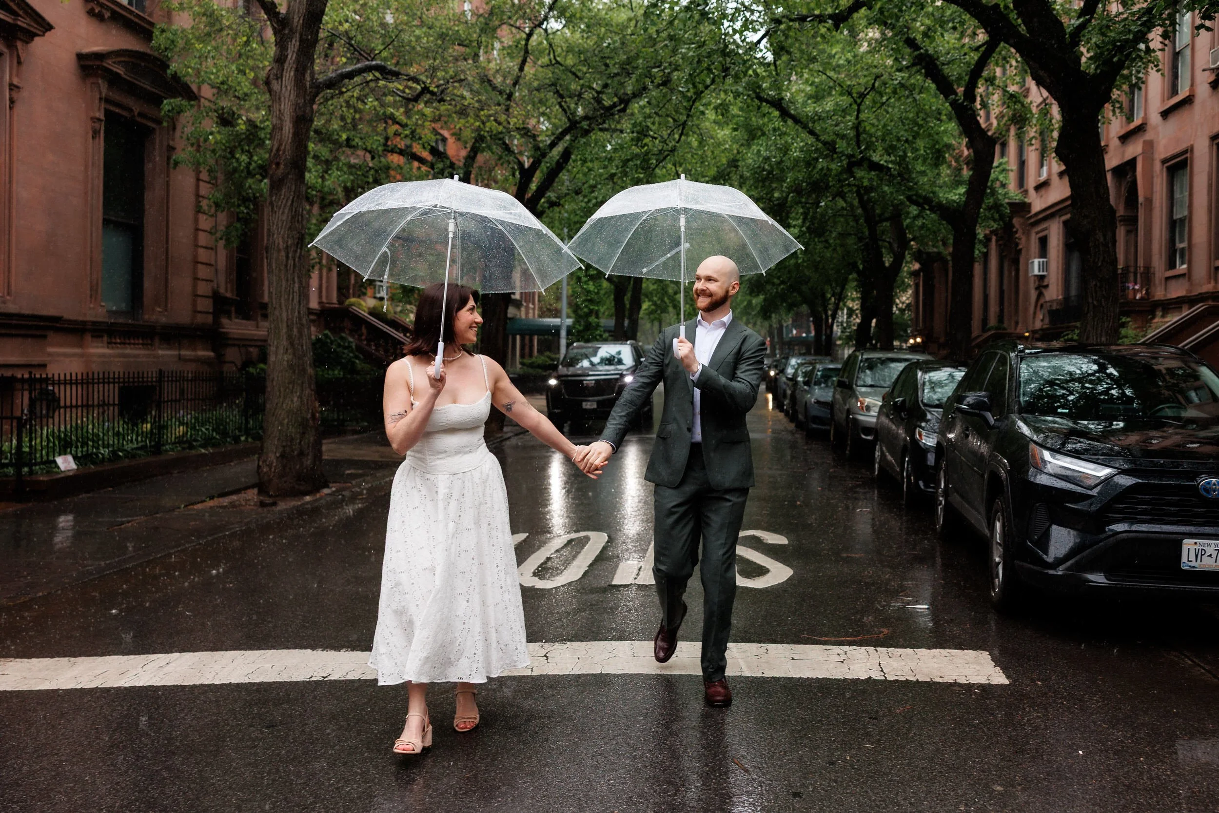A newlywed couple holding hands and walking down a street together as they each hold clear umbrellas 