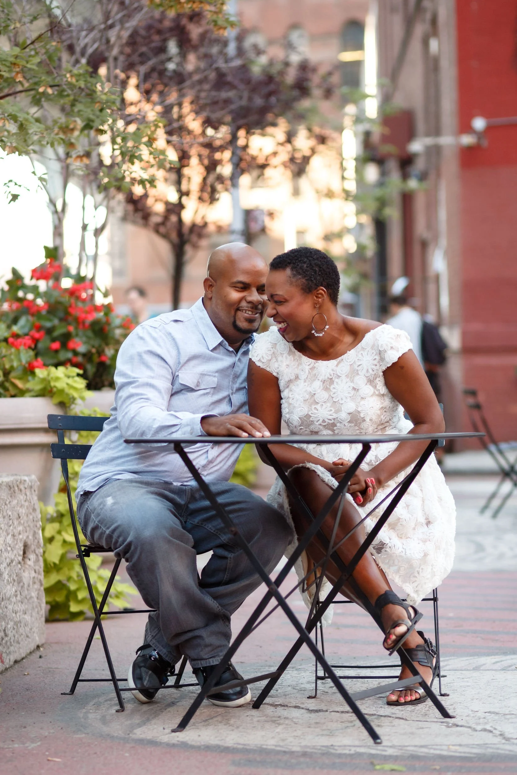 A couple sitting at a small outdoor table leaning into each other as they laugh 