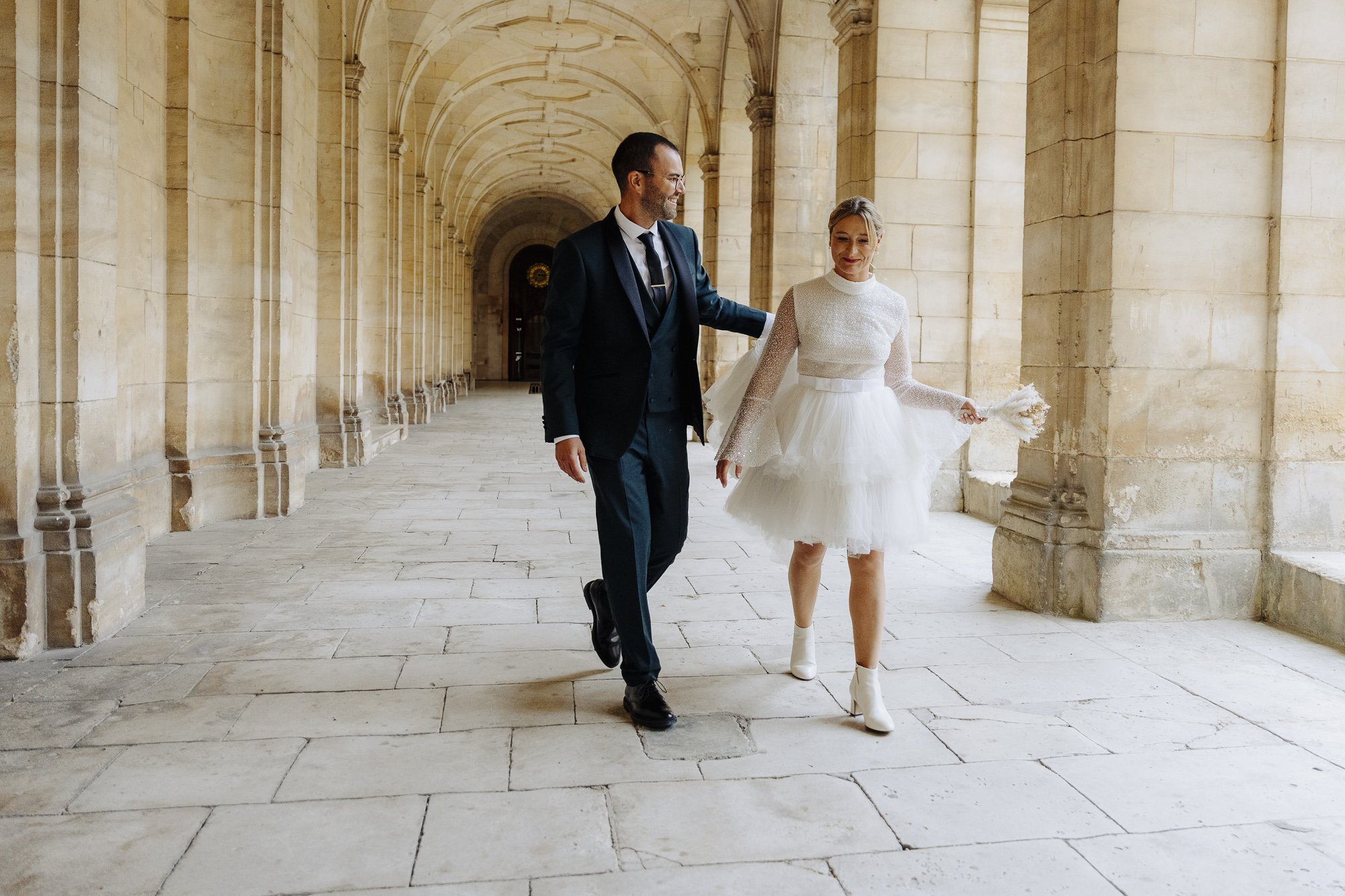 Un couple marchant dans un couloir voûté, la femme porte une robe de mariée courte et l'homme un costume.