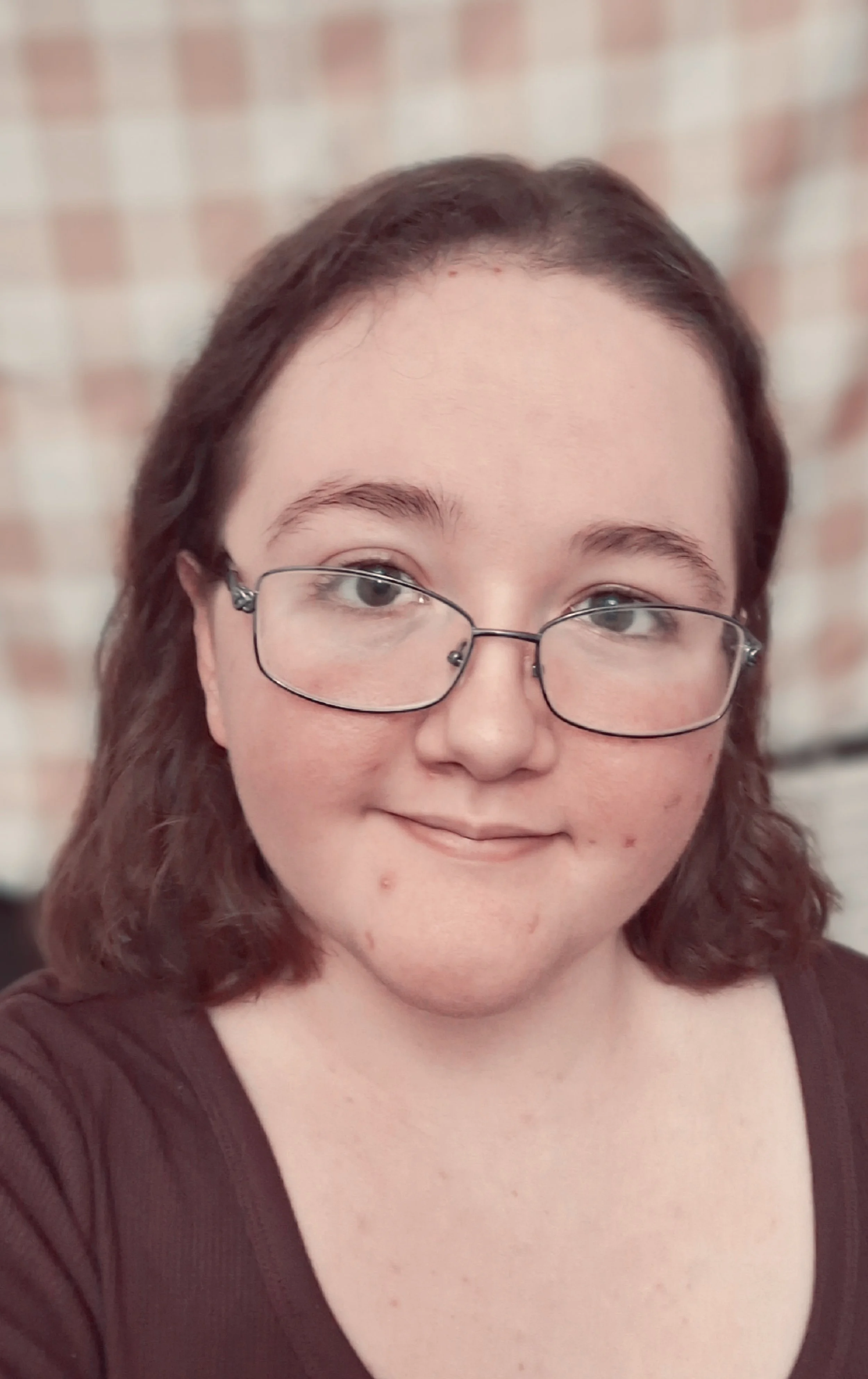 A young woman with glasses and shoulder-length brown hair smiling, with a brick wall background.