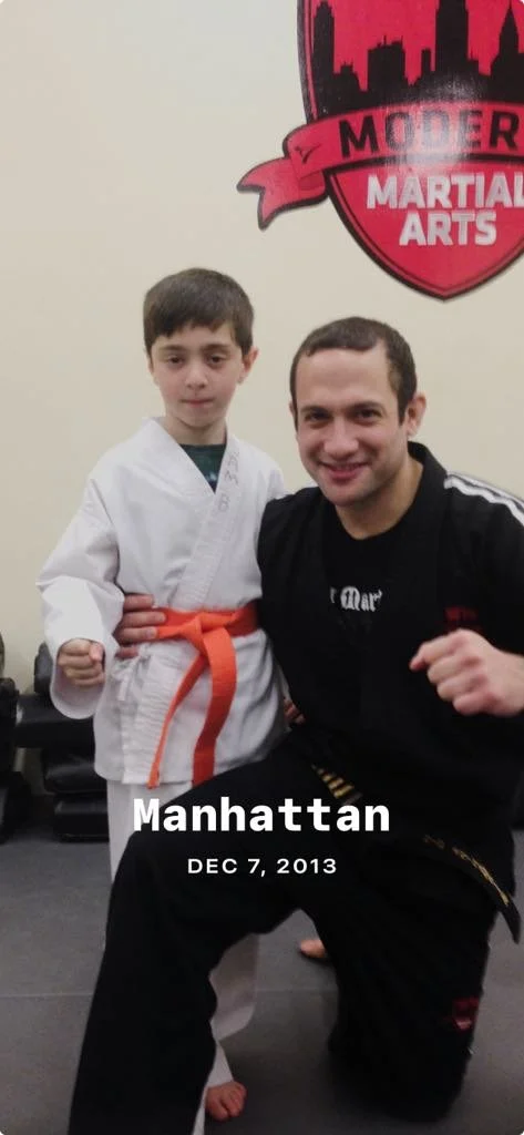 A child in a white martial arts uniform with an orange belt poses with an adult in a black uniform, standing in front of a Modern Martial Arts logo. The text "Manhattan DEC 7, 2013" is overlaid on the image.