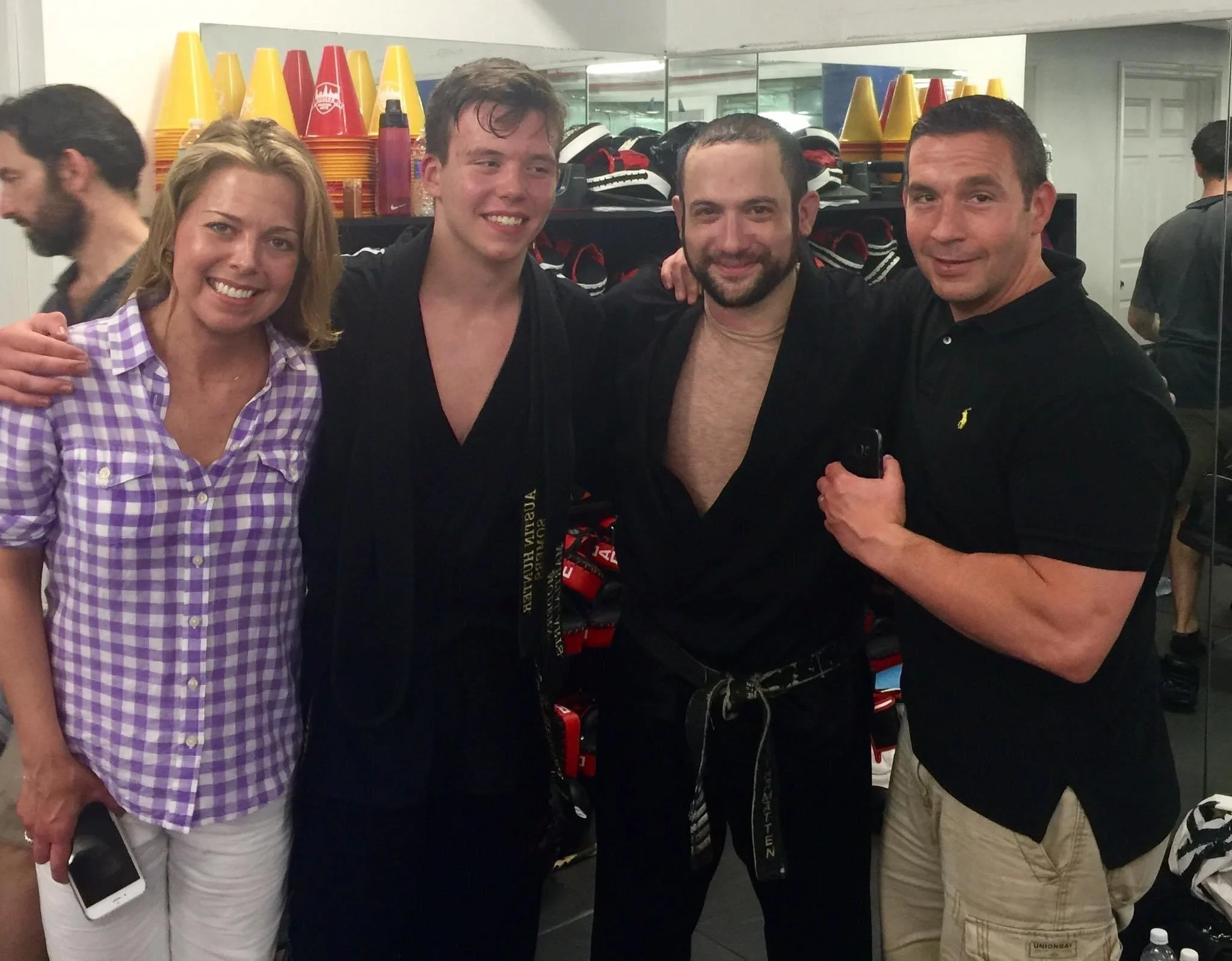 A group of four people smiling in a martial arts studio with gear and cones in the background. Two individuals are wearing martial arts attire while the other two are in casual clothes.