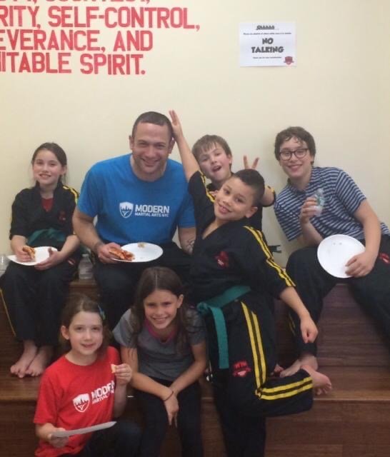 Group of kids with an adult in martial arts attire sitting and eating pizza, with wall text promoting positive values.