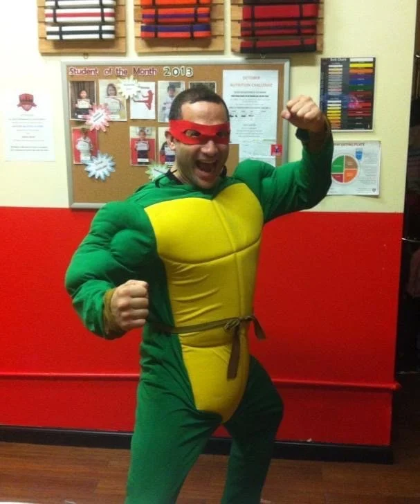 Person in green turtle costume with red eye mask posing energetically indoors with a bulletin board in the background.