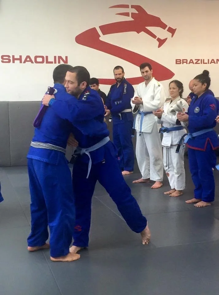 Two people in blue Brazilian jiu-jitsu gi practicing a hold, surrounded by others in martial arts uniforms at a training facility with 'Shaolin' and 'Brazilian' on the wall.