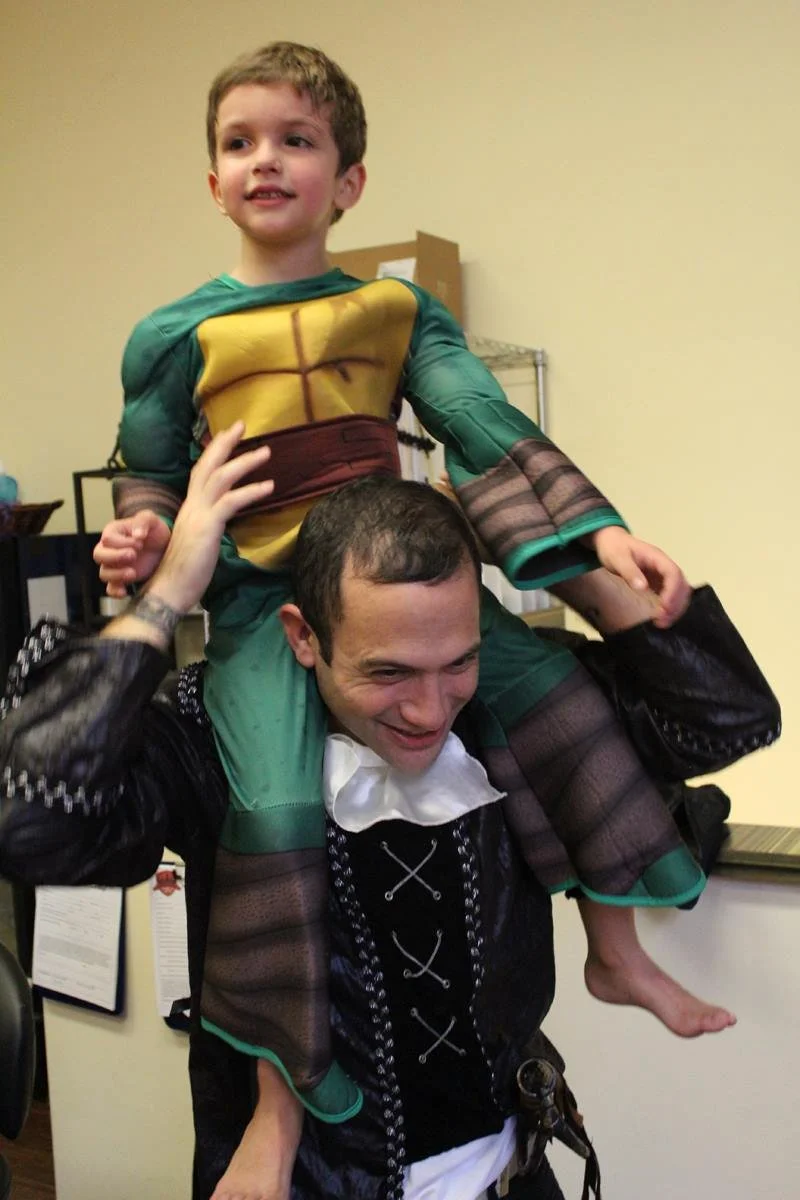 Child in Ninja Turtle Halloween costume, sitting on Mr. A's shoulders, both smiling, indoors.