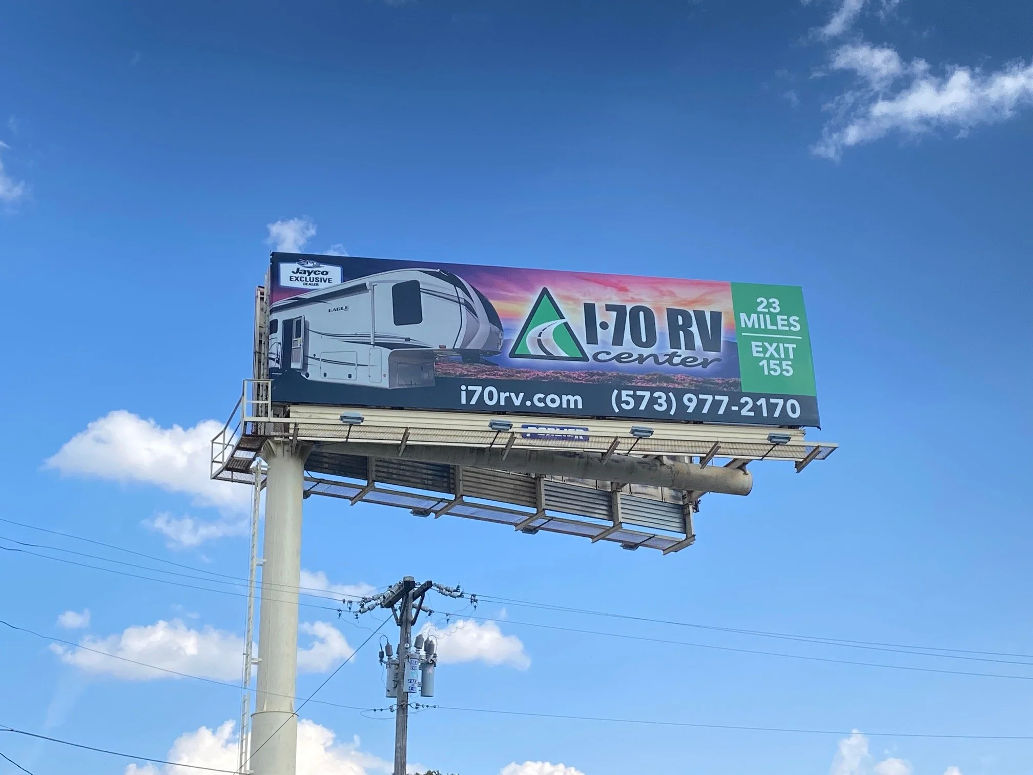 Billboard advertising i70 RV Center with a graphic of a recreational vehicle, information about location at 23 miles and exit 155, contact number, and website, against a blue sky with clouds.