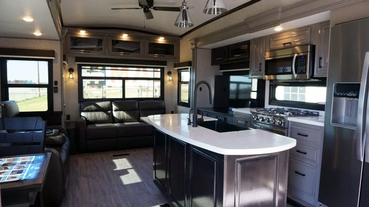 Interior of a modern RV with a kitchen island, stainless steel appliances, a TV, and a living area with a black sofa by windows.