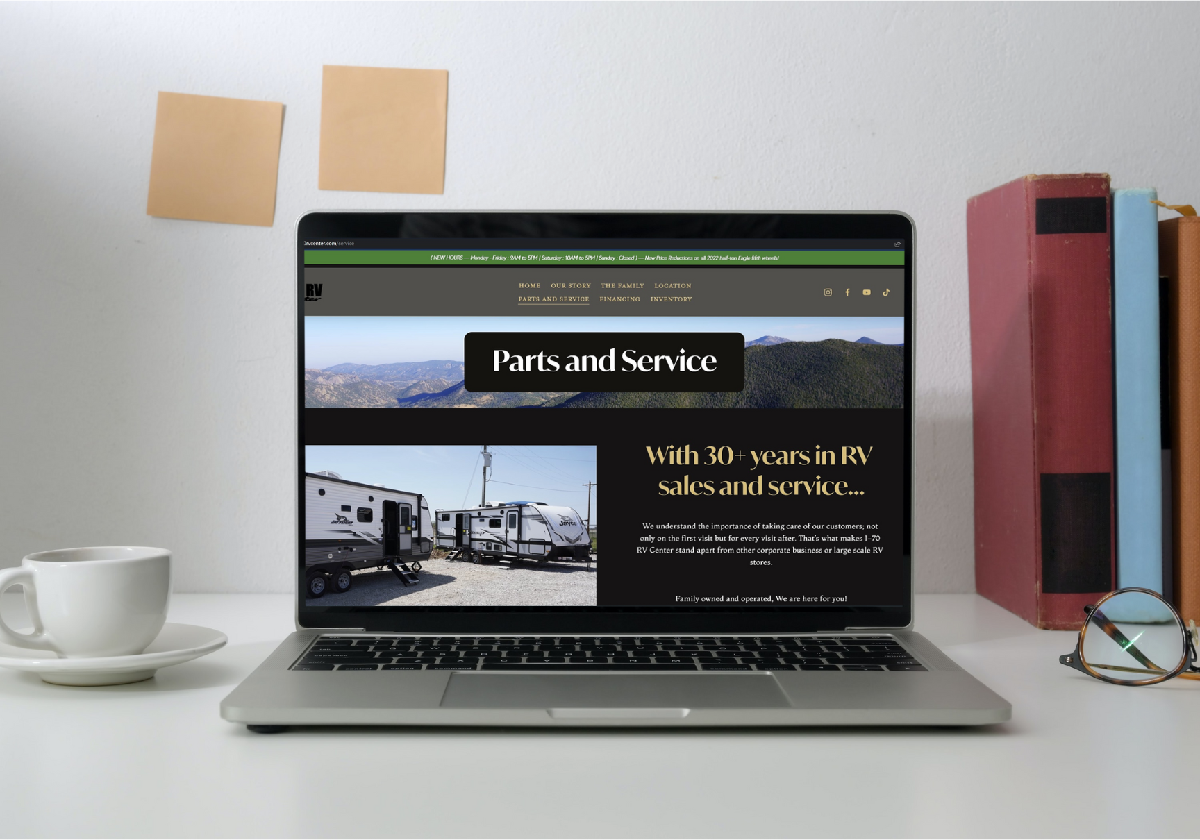 Laptop on a white desk displaying an RV parts and service website with an image of two travel trailers and mountain scenery. Surrounding objects include a white coffee cup with a saucer, a pair of glasses, and three vertically stacked books with red, blue, and orange covers. Two beige sticky notes are attached to the white wall behind the desk.