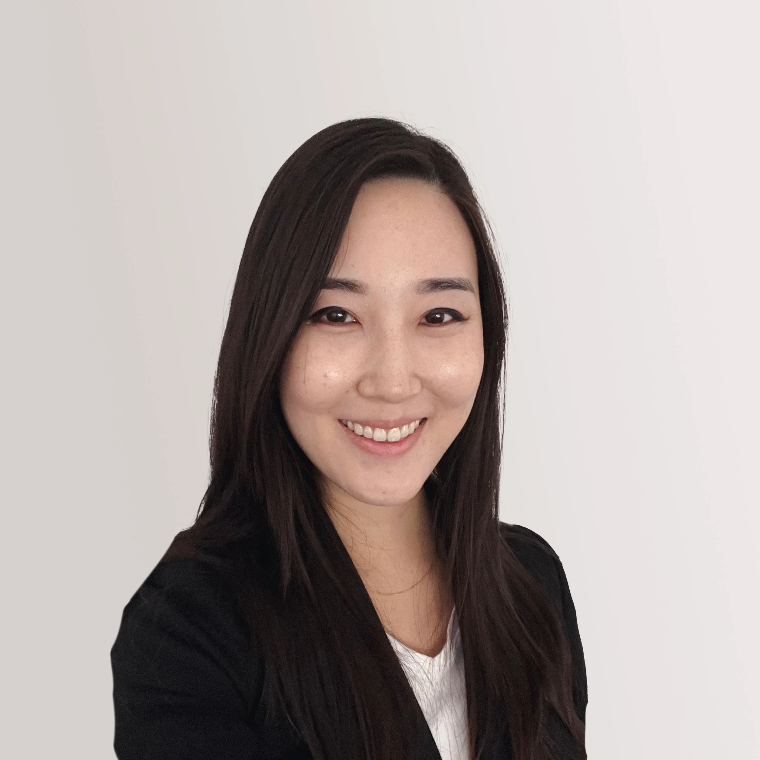 A young woman with long dark hair, smiling, wearing a black blazer and a white top, standing against a plain white background.