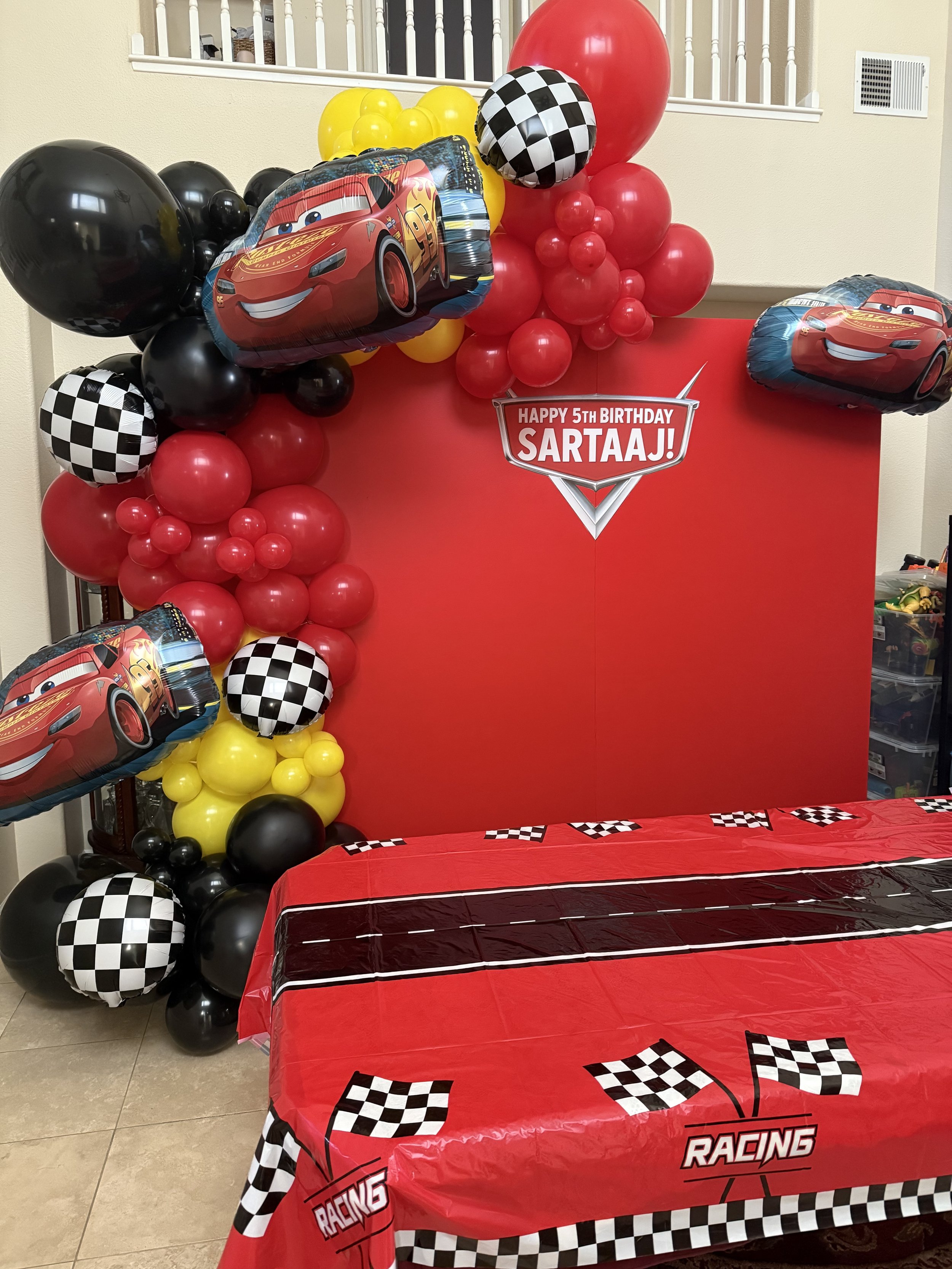 Decorations for a 5th birthday party with a racing car theme. Red backdrop with balloons in black, yellow, and checkered patterns. Mylar balloons featuring Lightning McQueen from Cars. Table covered with a red racing-themed tablecloth with checkered flags.