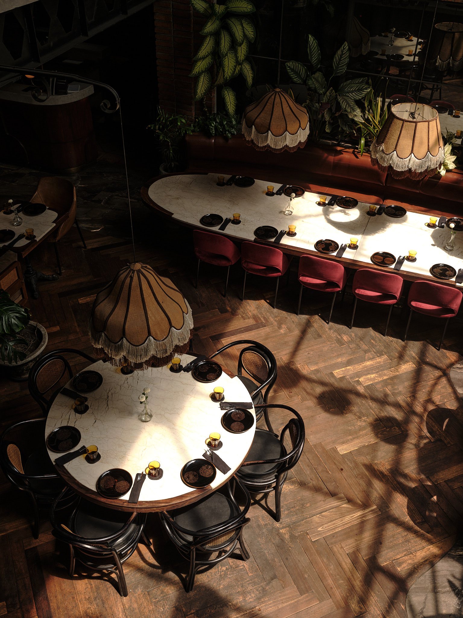 A top-down view of a restaurant dining area with six round white and marble tables, each set with black plates, yellow glasses, and black utensils. There are black chairs around the tables, and decorative lamps with scalloped beige shades hang above. Green plants are visible along the back wall, and a wooden floor completes the scene.