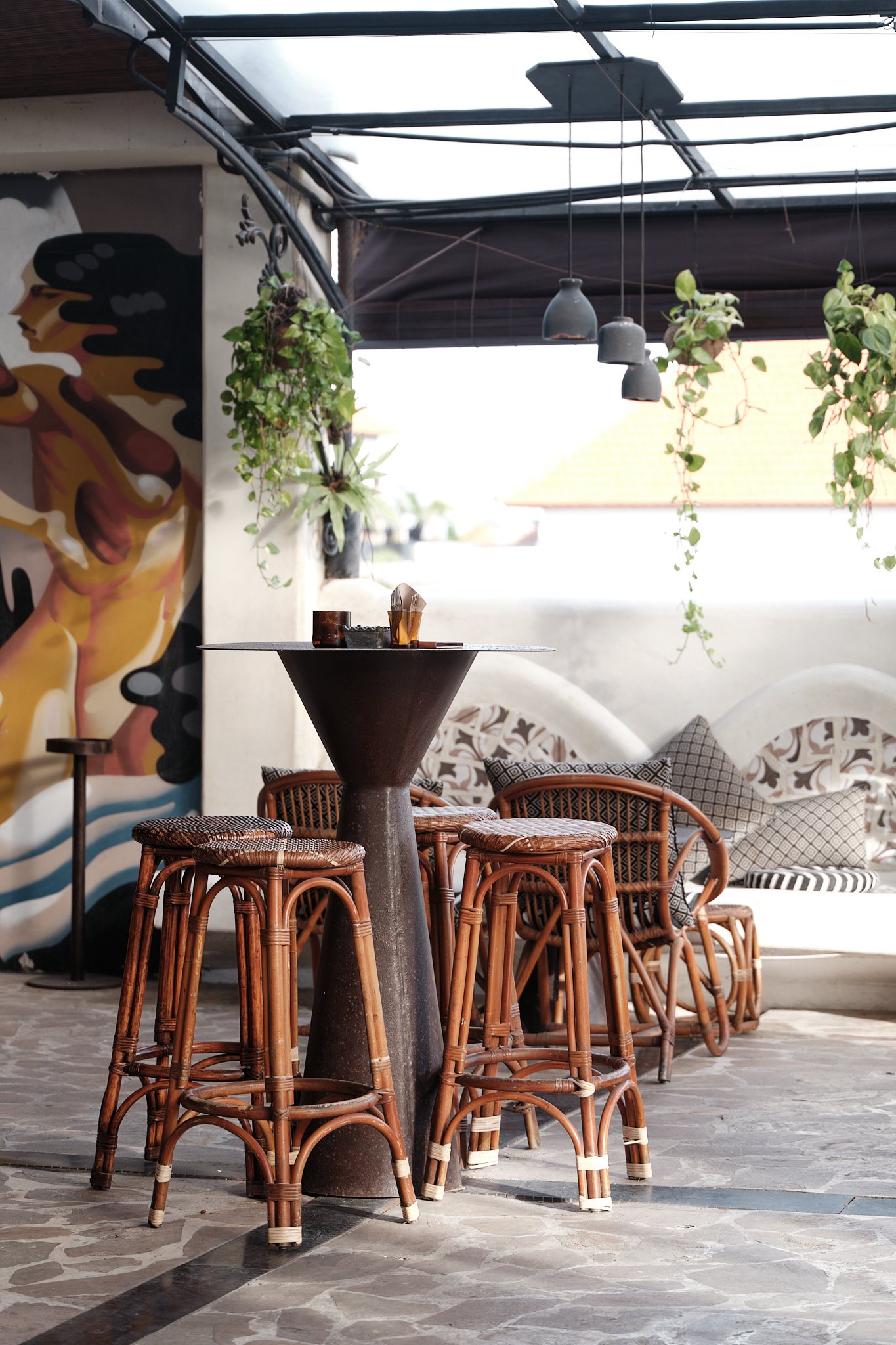 Indoor seating area with woven rattan chairs surrounding a round black table, hanging pendant lights, and hanging plants, with a mural partially visible on the wall and natural light coming from outside.